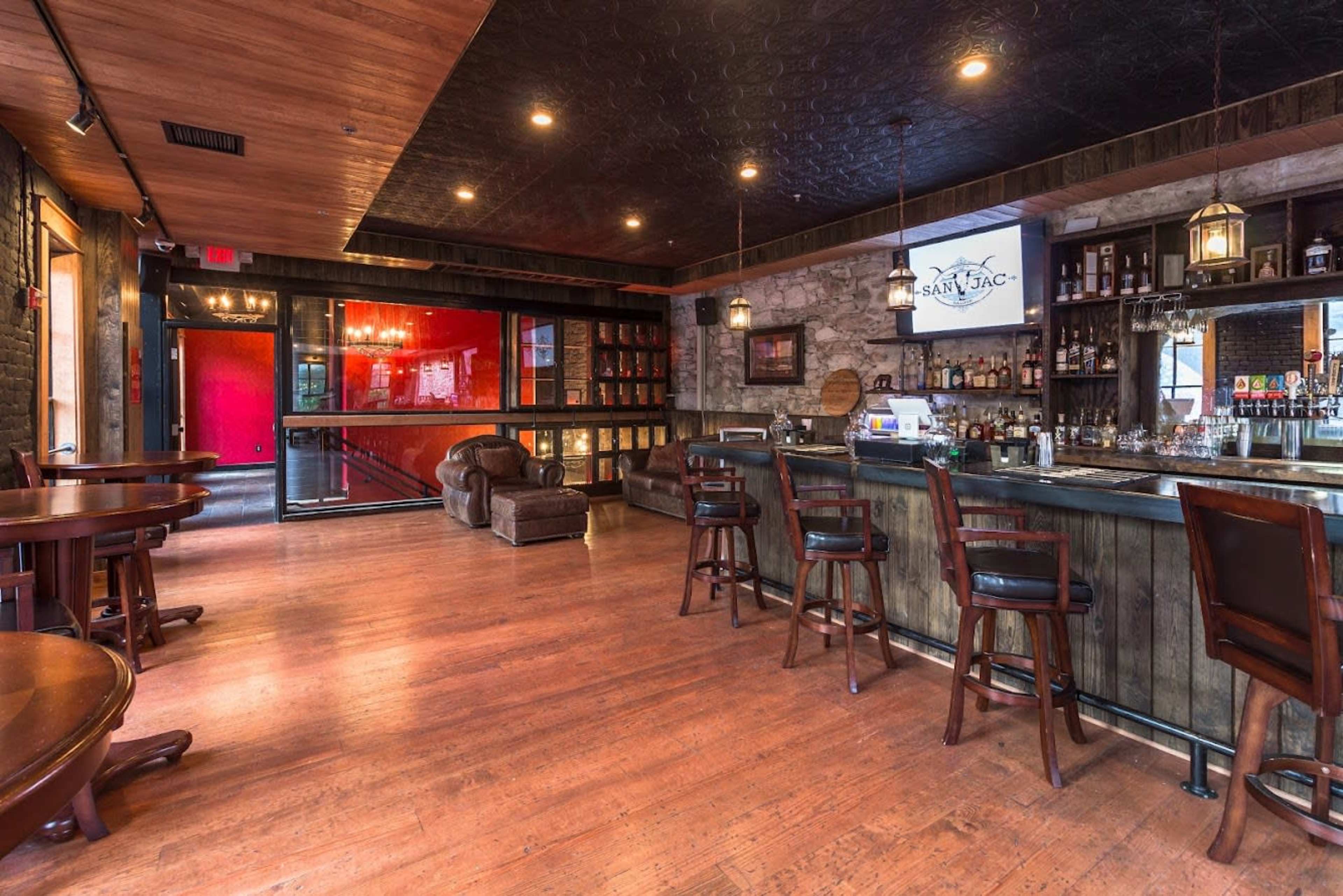 A spacious bar area with wooden floors, a long bar counter, and several seating arrangements, illuminated by overhead lights.