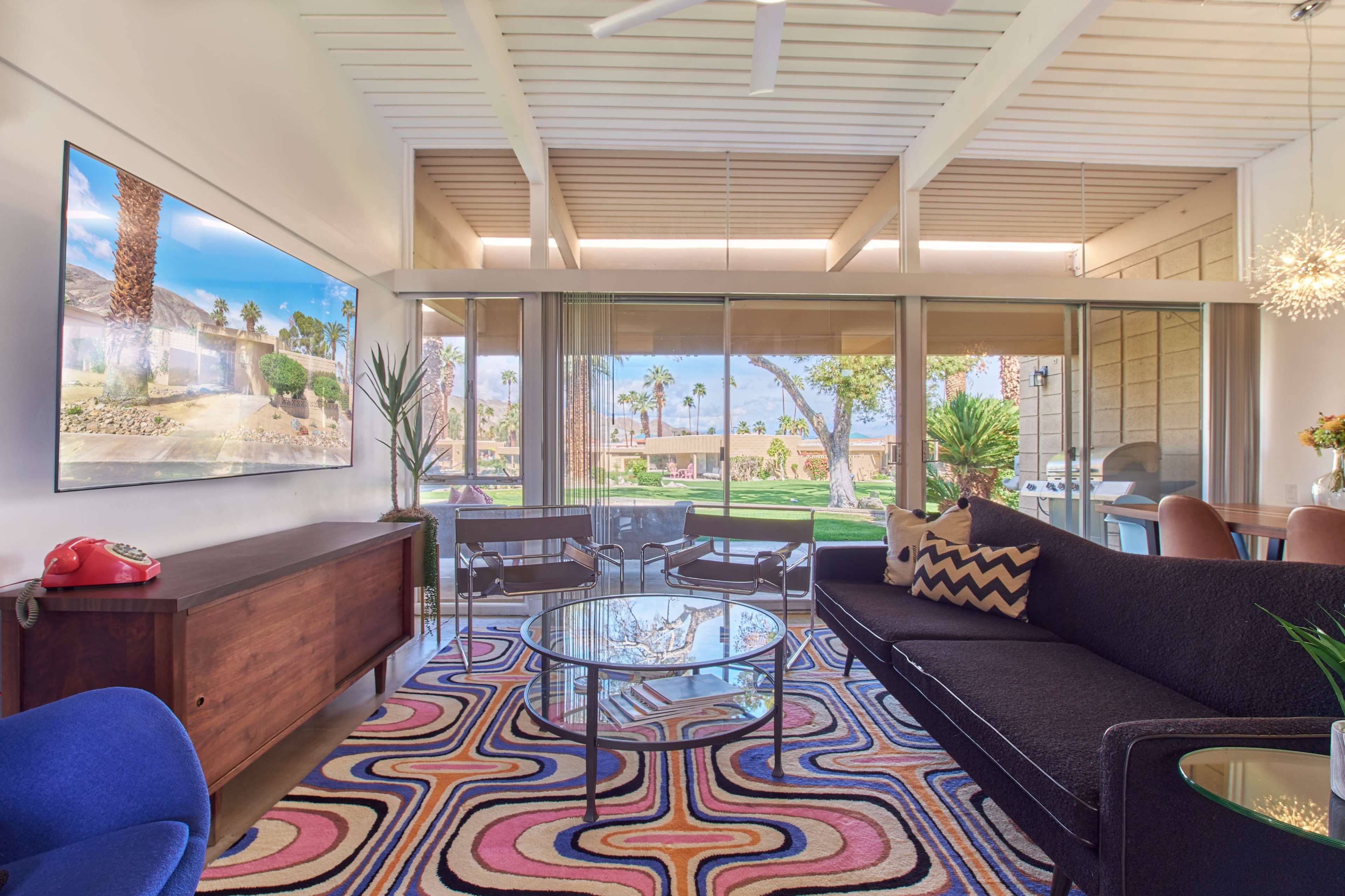 The image shows a modern living room with a large window overlooking a landscaped outdoor area, featuring a black sofa, colorful rug, and sleek furniture.