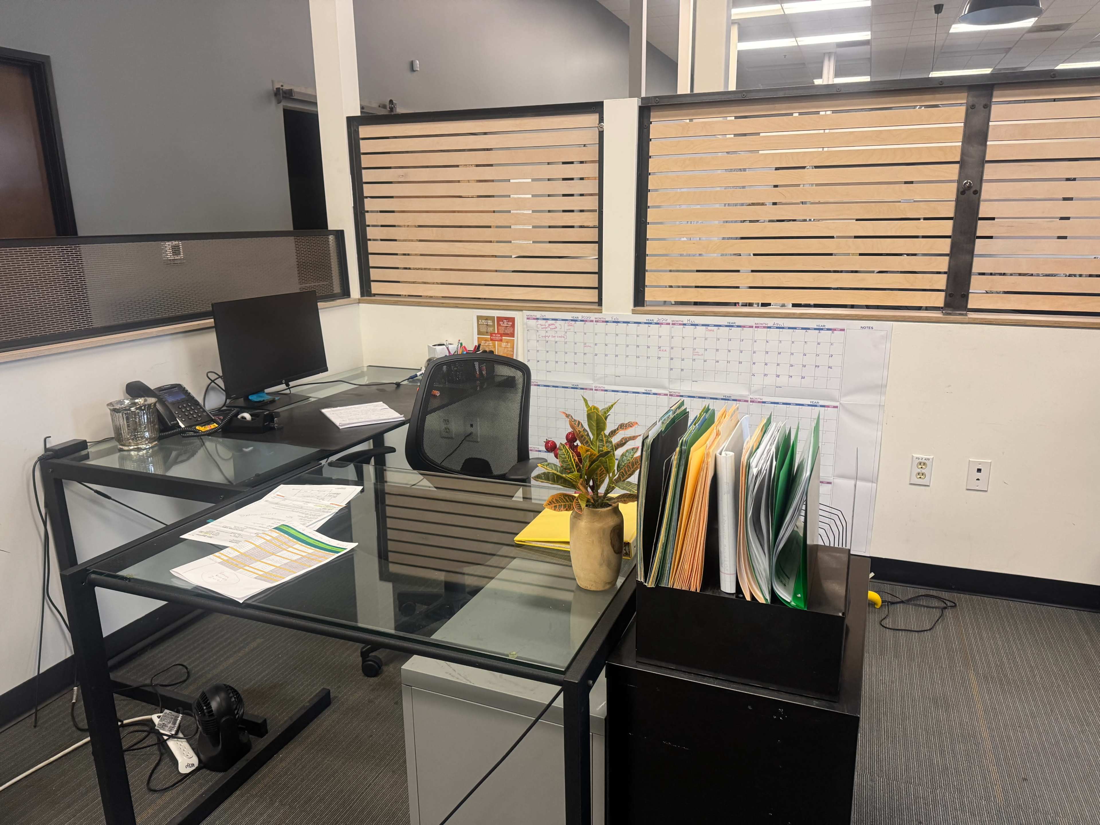 The image shows an office space featuring a desk with a computer, telephone, organized files, and a small decorative plant.