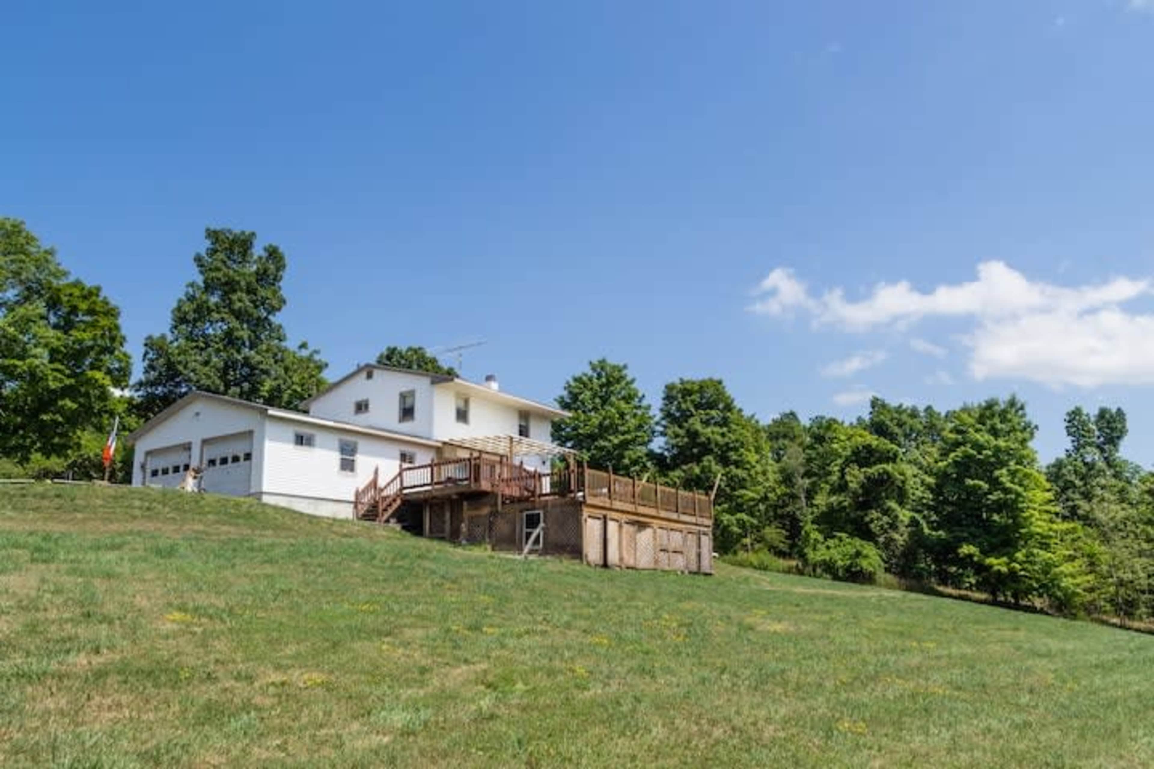 A two-story white house with a wooden deck and garage sits on a grassy hillside surrounded by trees.