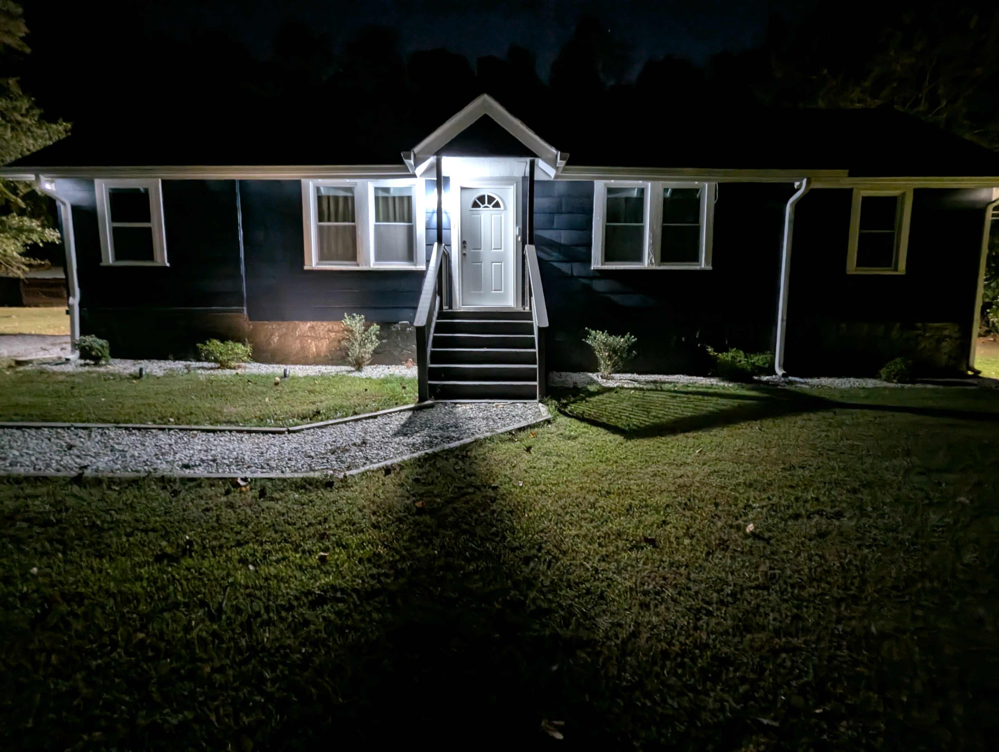 A dark-clad house is illuminated by a porch light at night, featuring a front staircase and a well-maintained lawn with gravel pathways.