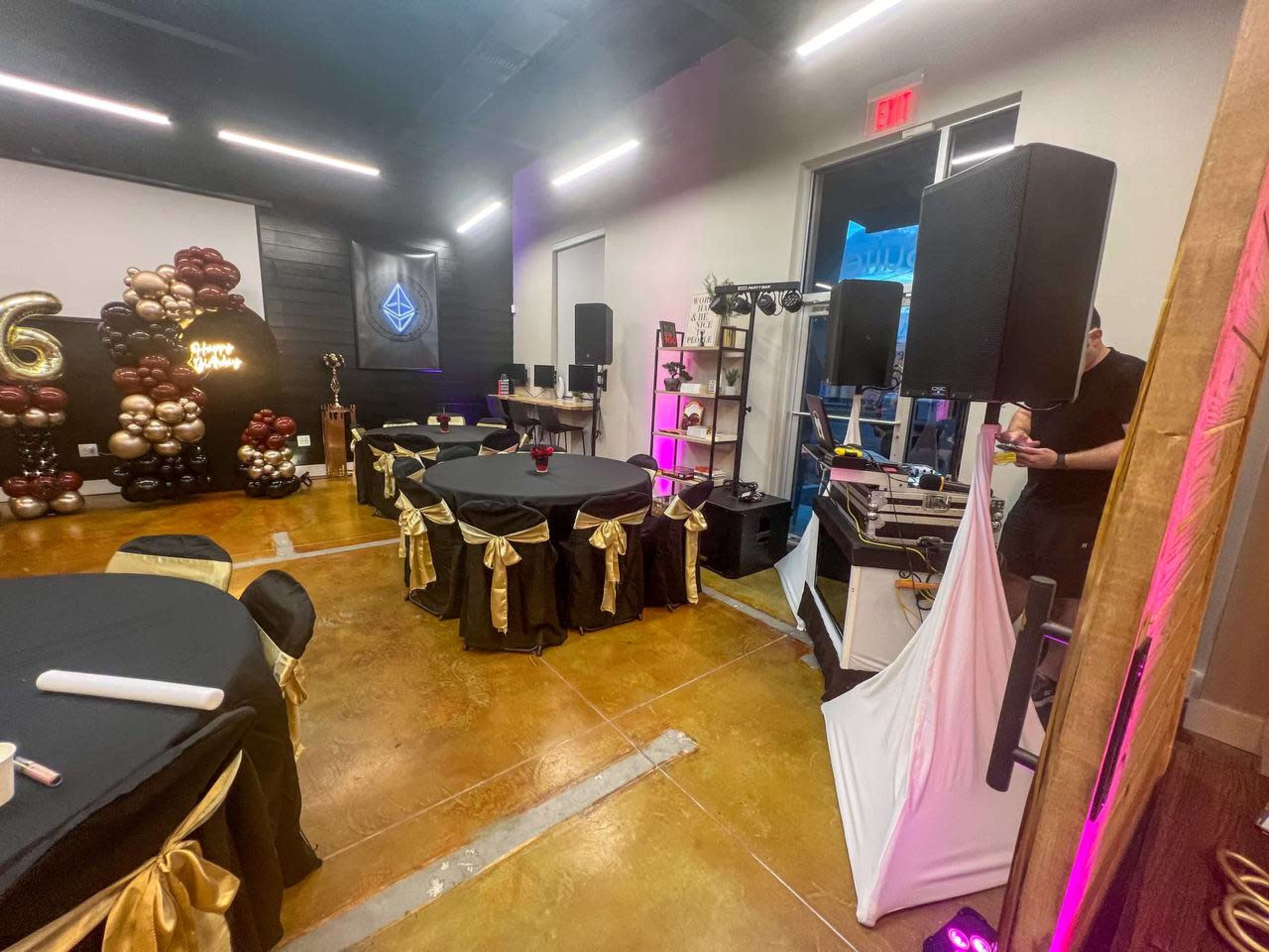 A party setup with black tables adorned with gold ribbons, a DJ booth, and decorative balloons in the background.