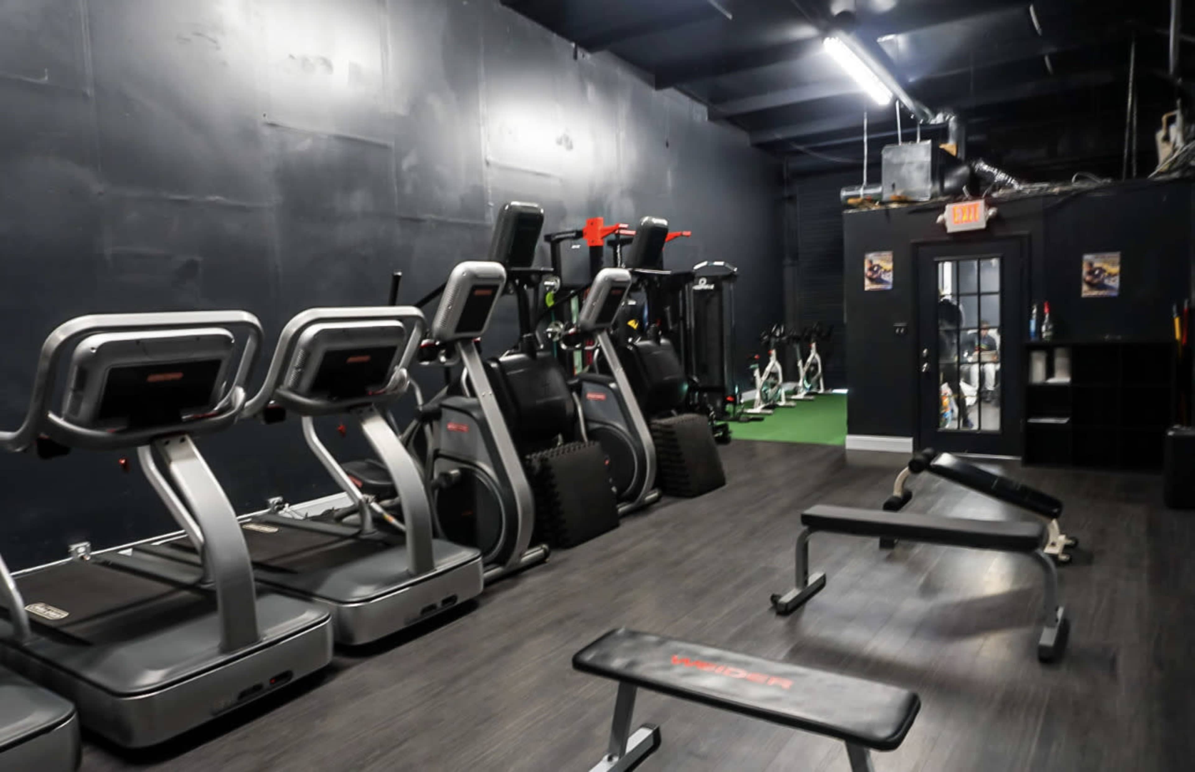 A gym interior with several exercise machines, including treadmills and benches, arranged along dark walls.