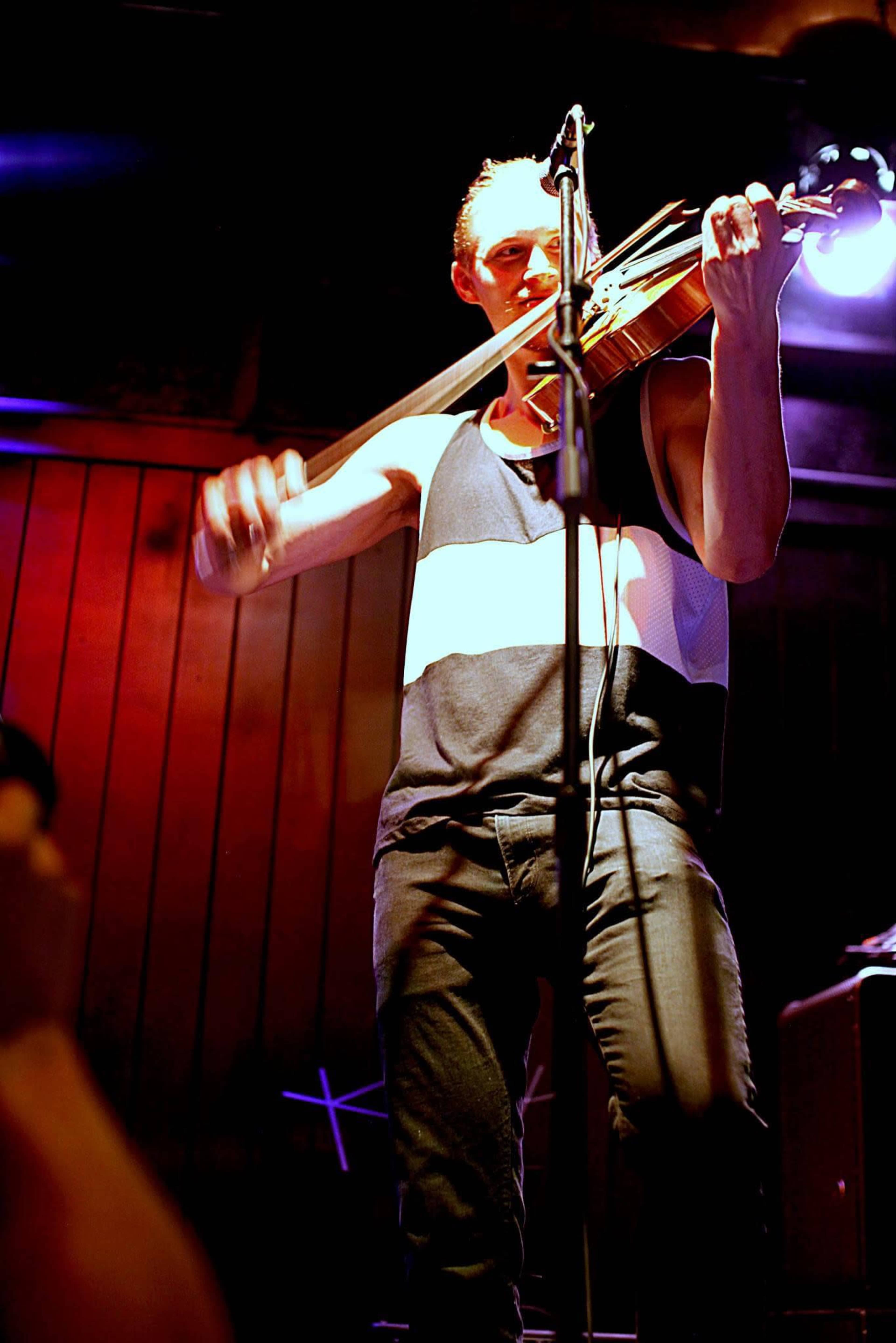 A musician plays the violin on stage under dim lighting, with a microphone in front of him.