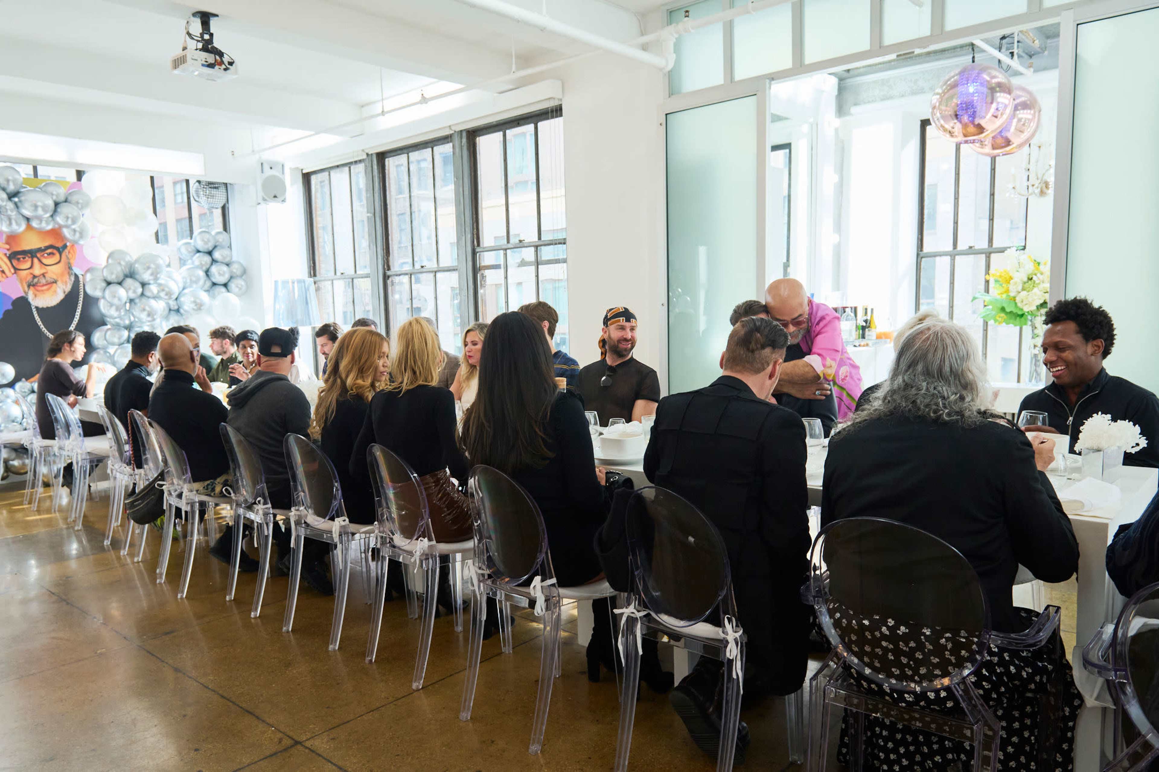 A long table is set for a gathering of people dressed in mostly black, with a large mural featuring a man with glasses in the background.