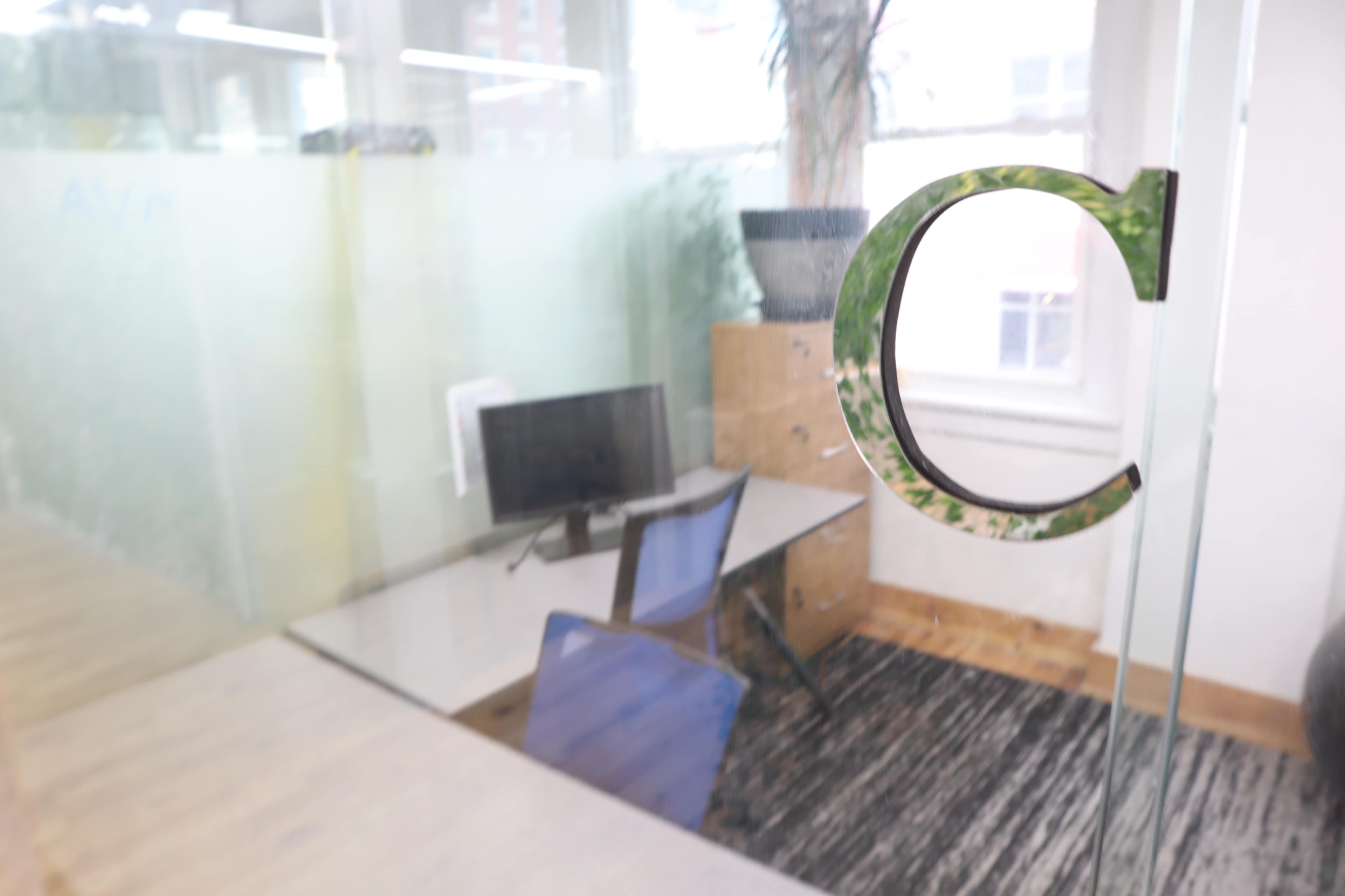 The image shows a modern office space with a glass wall featuring the letter "C," a desk with a computer, and a potted plant in the corner.