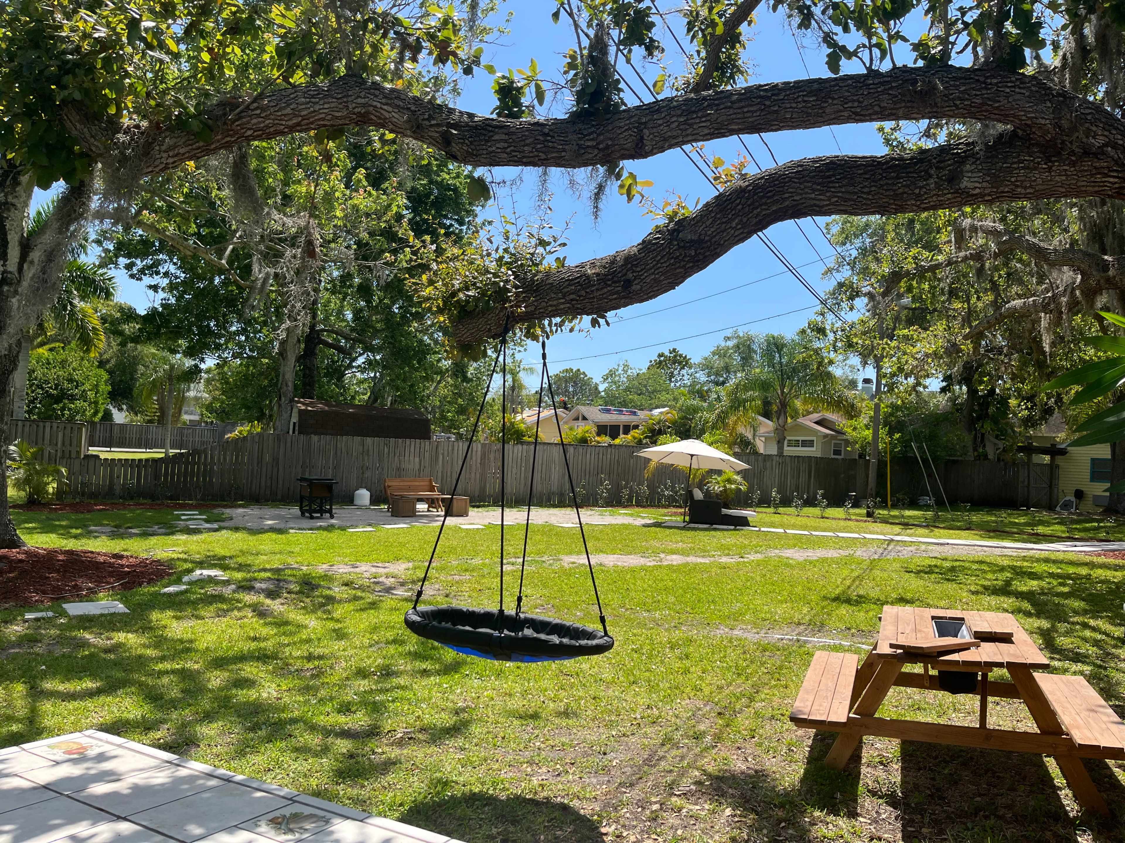 A swing hangs from a tree branch in a grassy backyard with picnic tables and an umbrella in the distance.