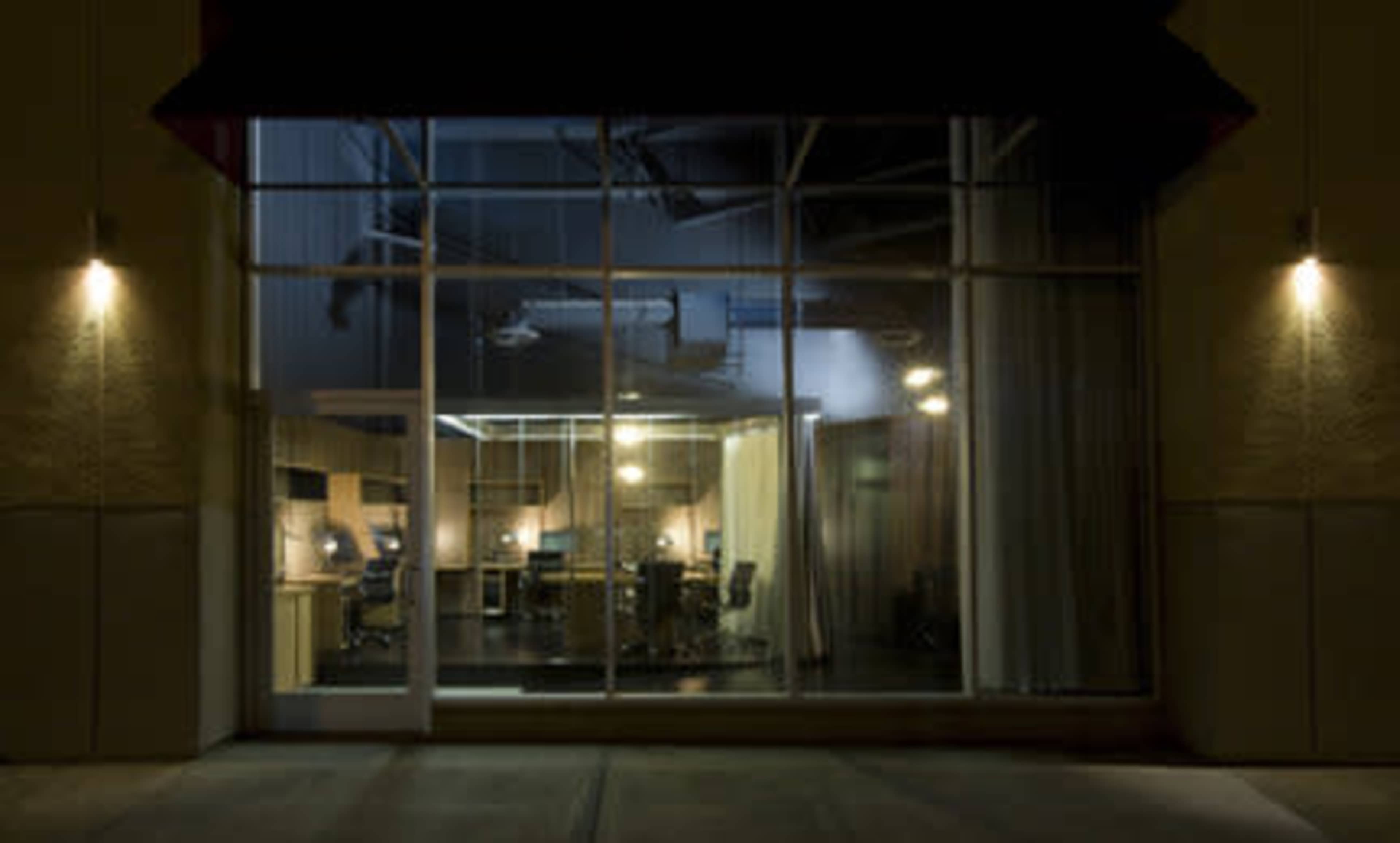 A dimly lit office space visible through large glass windows, with multiple workstations and chairs arranged inside.