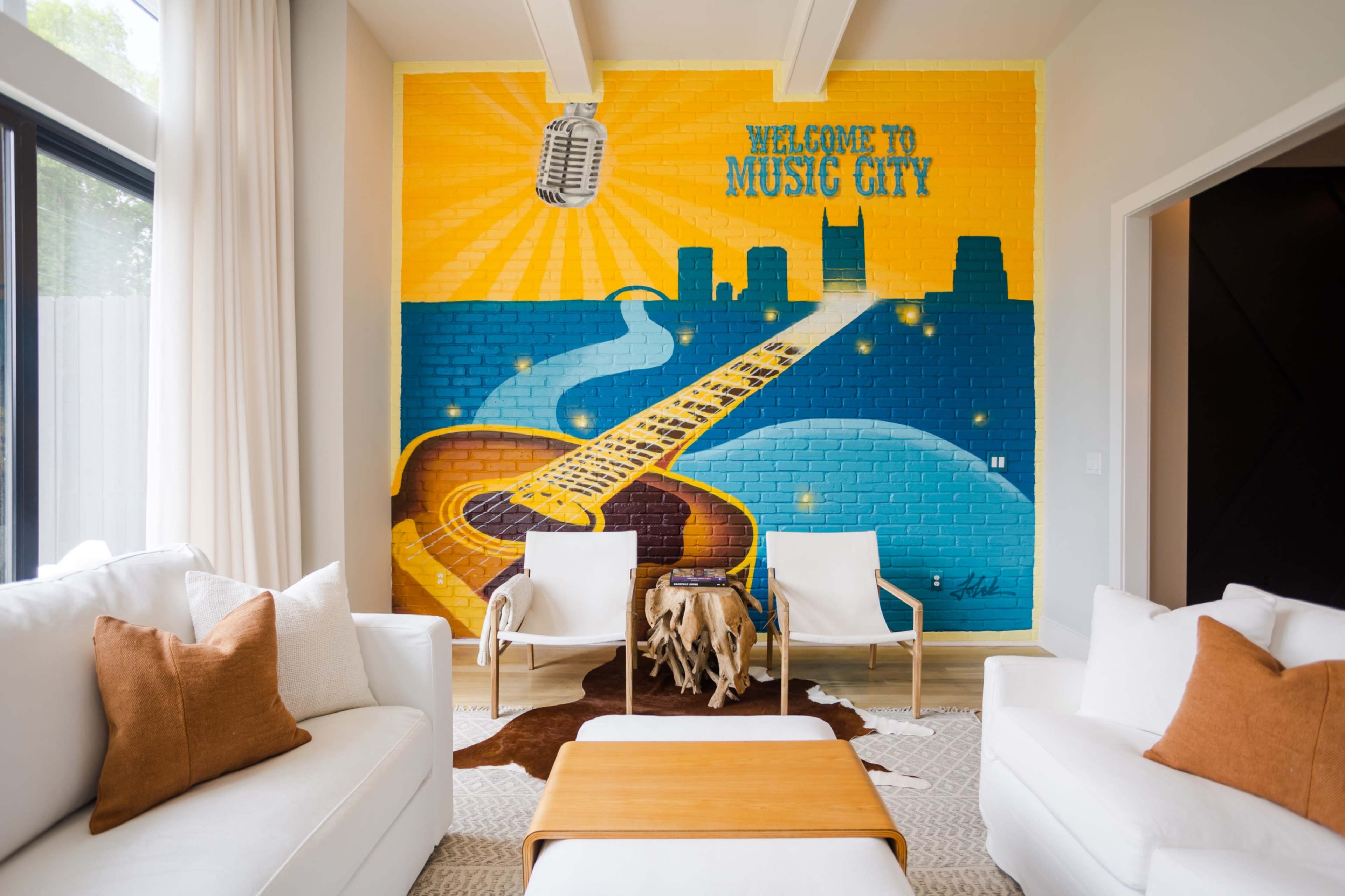 The image features a vibrant mural depicting a guitar leading to a city skyline, with the phrase "Welcome to Music City" prominently displayed, in a modern room furnished with white sofas and a wooden coffee table.