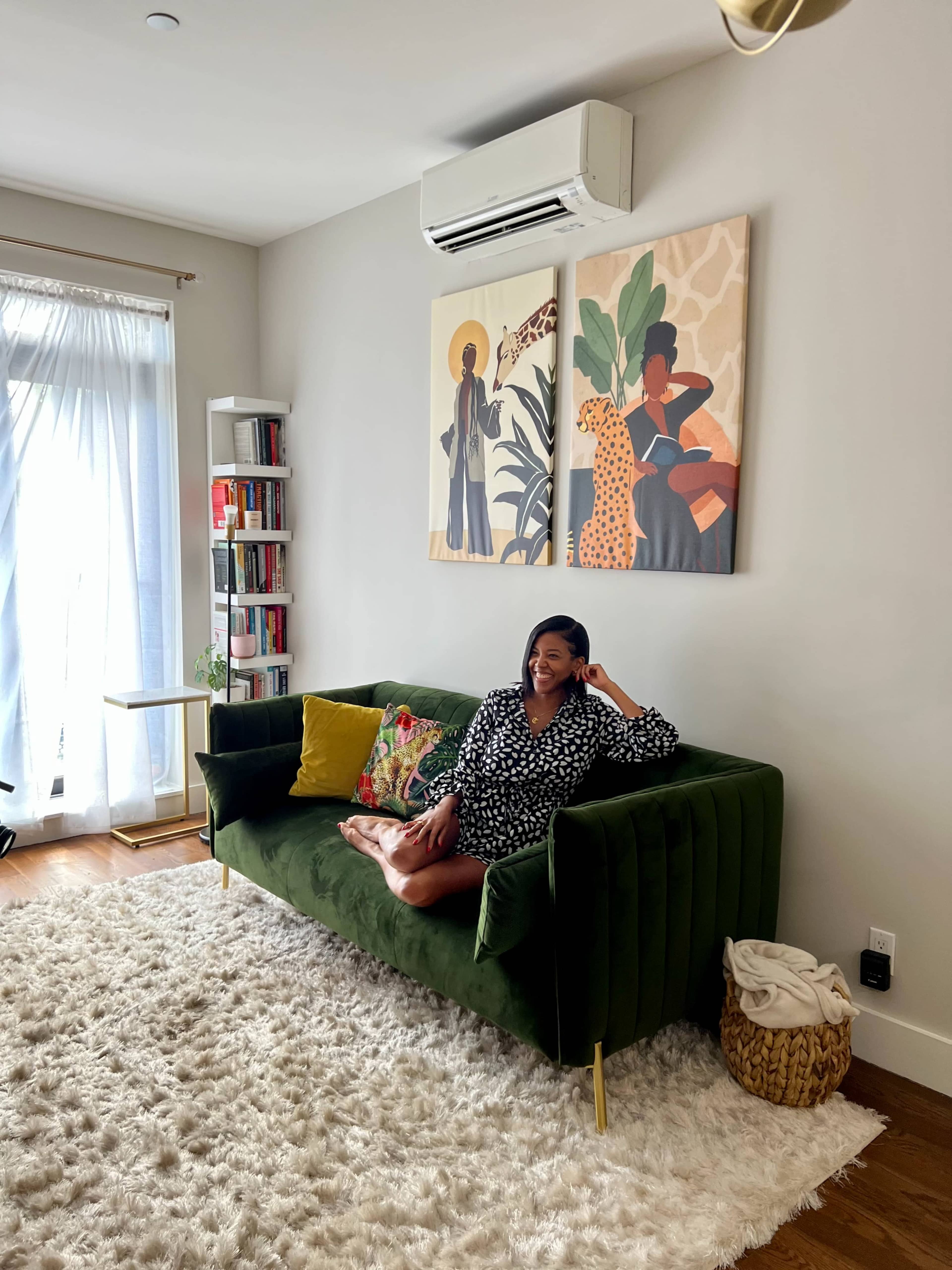 A person sits on a green sofa in a bright living room, decorated with art and a bookshelf, with sunlight streaming through a sheer curtain.