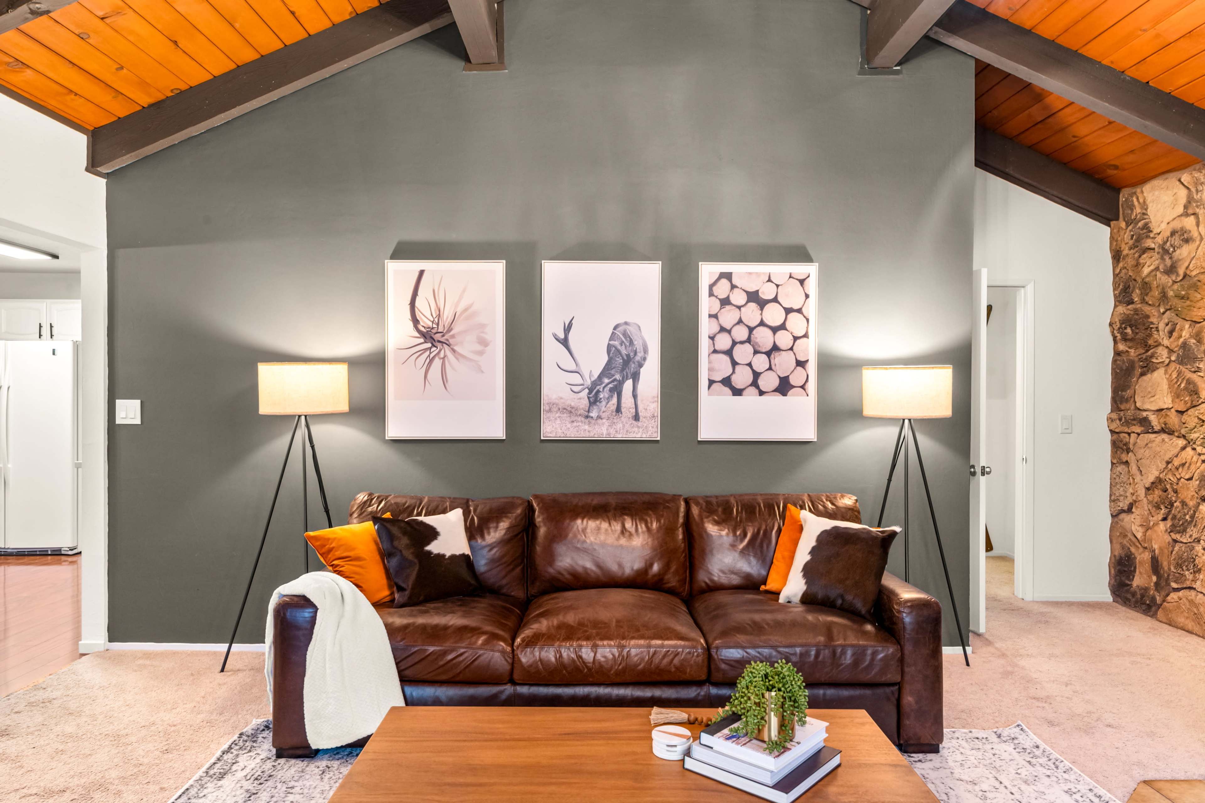 A leather sofa with accent pillows sits in a living room featuring three framed artworks on a gray wall and two floor lamps beside it.