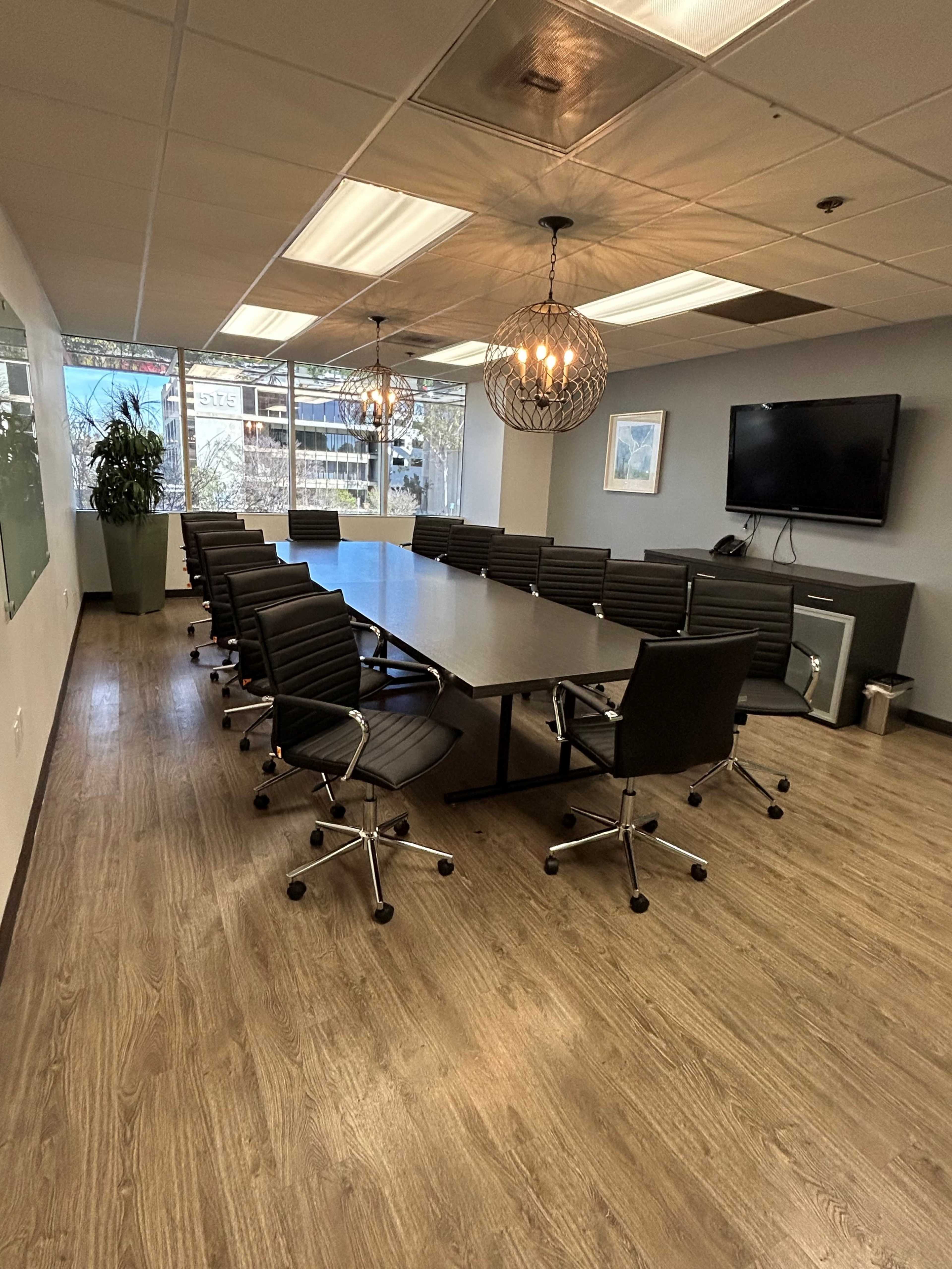 A modern conference room features a long table surrounded by ergonomic chairs, with large windows providing natural light and showcasing greenery outside.