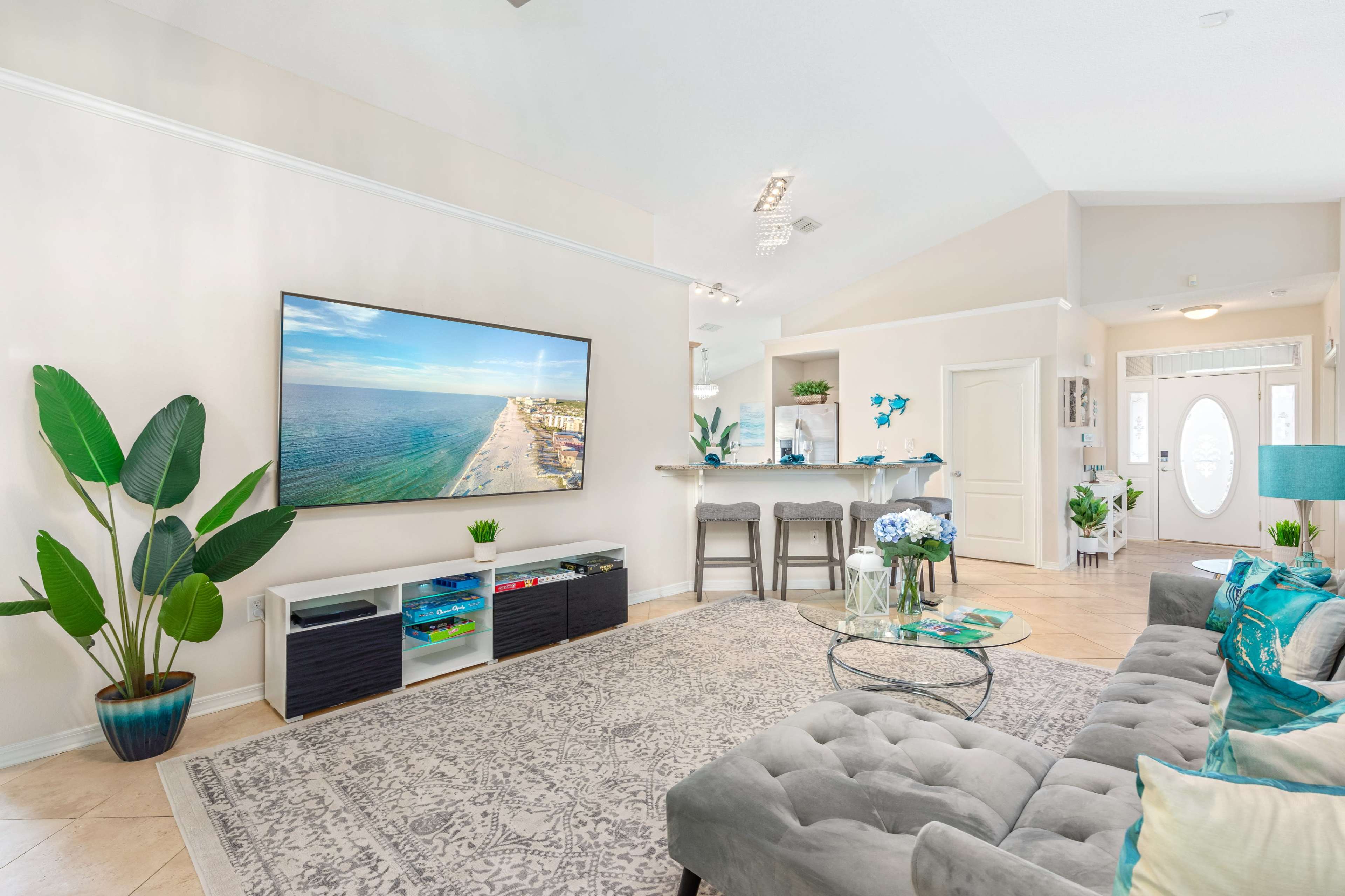 The living room features a large flat-screen TV mounted on the wall, a gray sofa, potted plants, and a light-colored rug, with a view of a bar area and front door in the background.