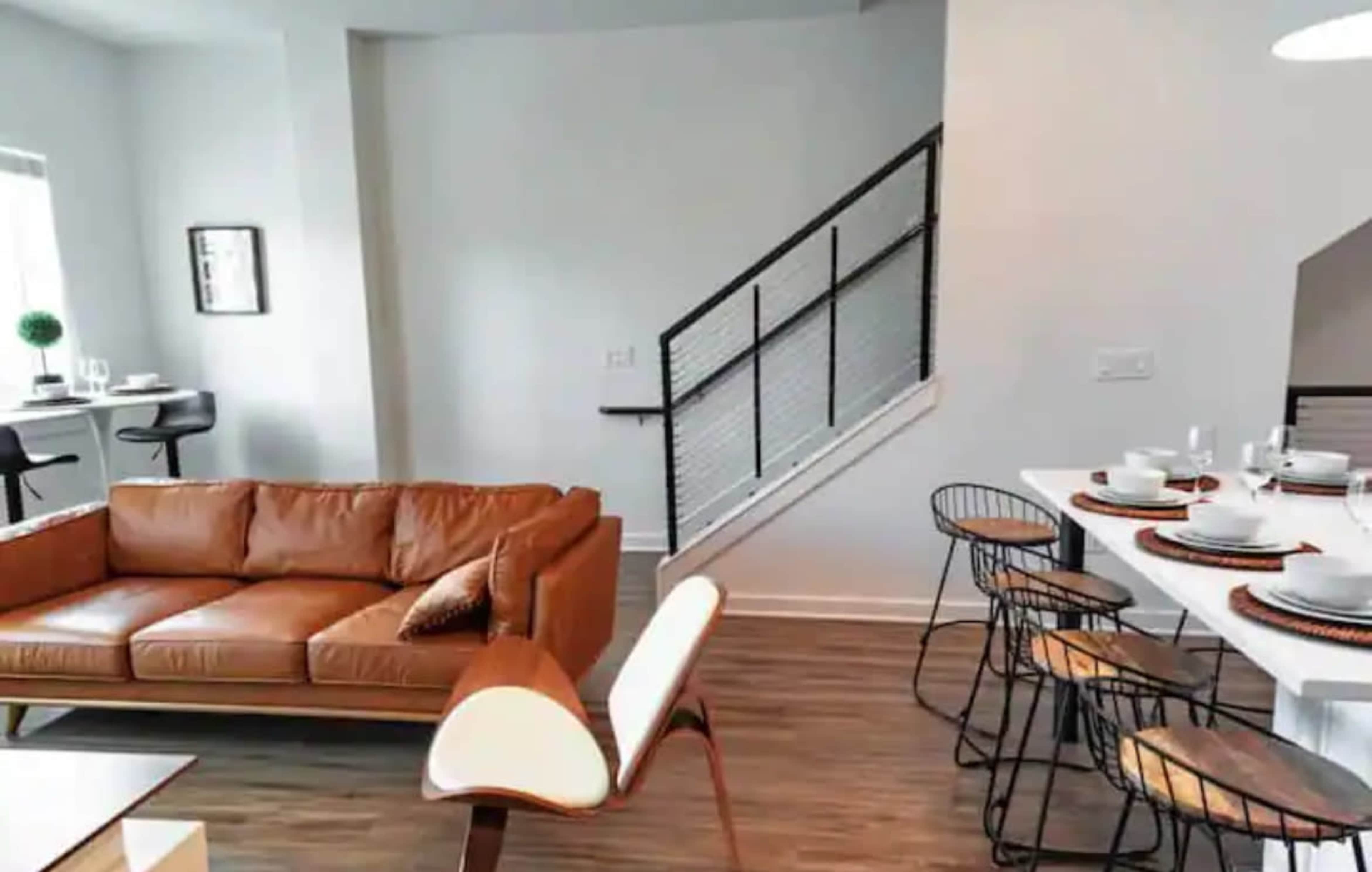 A modern living space featuring a brown leather sofa, a dining table set for six, and a staircase leading to an upper level.