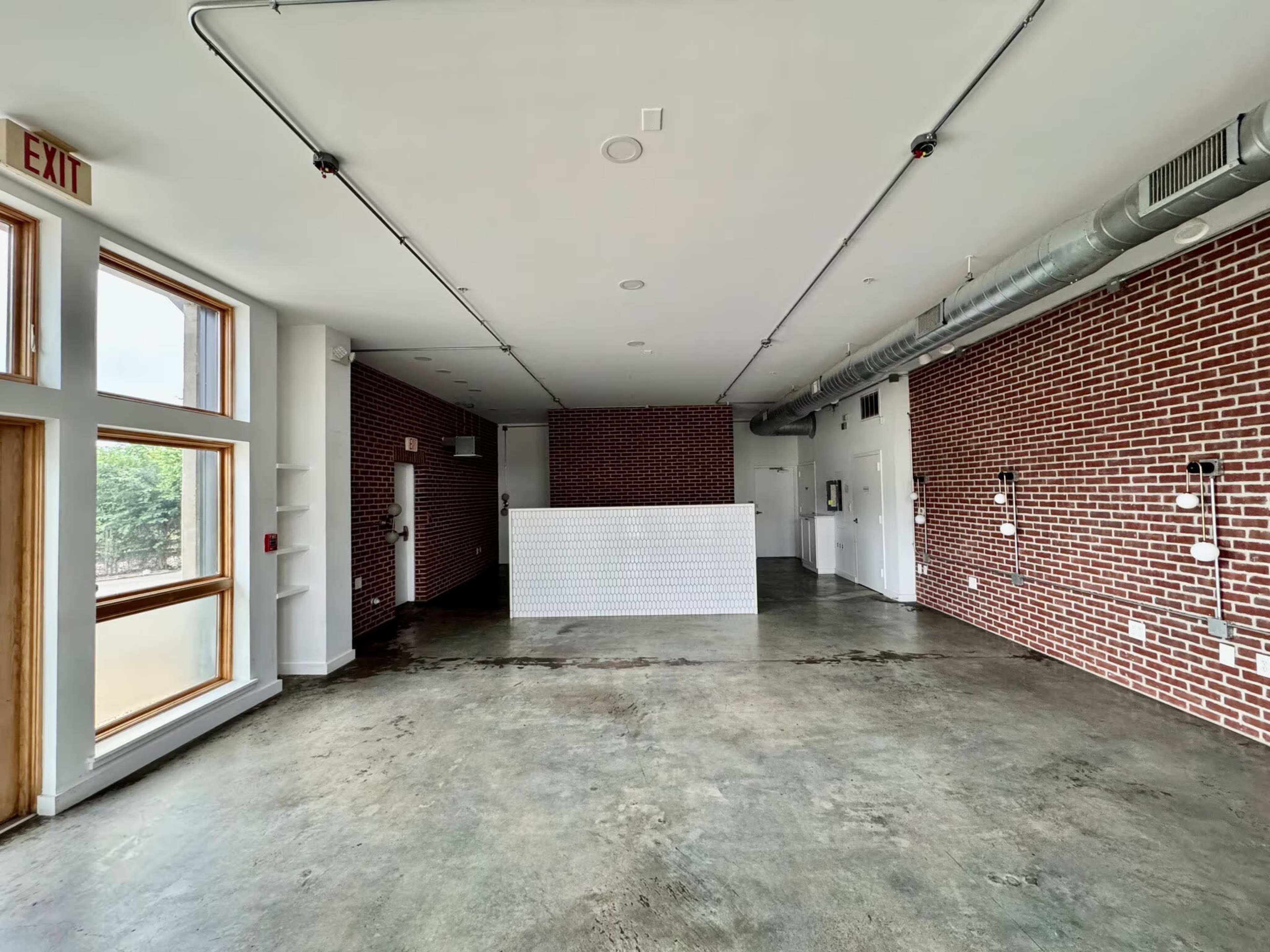The image shows an empty interior space with concrete flooring, exposed brick walls, and a white reception desk positioned in the center.