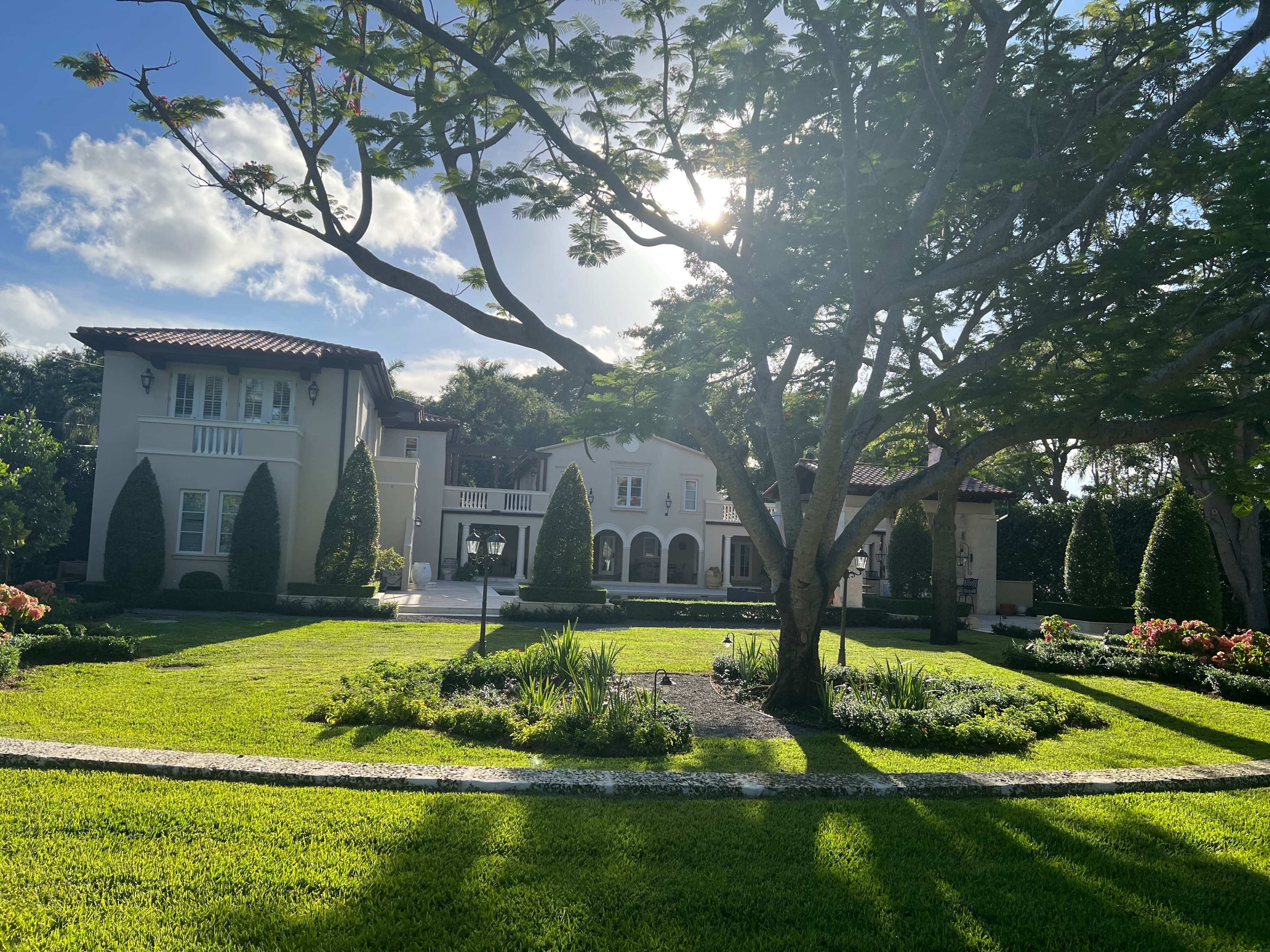 A large, elegant house with Mediterranean architectural features is surrounded by manicured lawns and neatly trimmed shrubs beneath a bright sky.