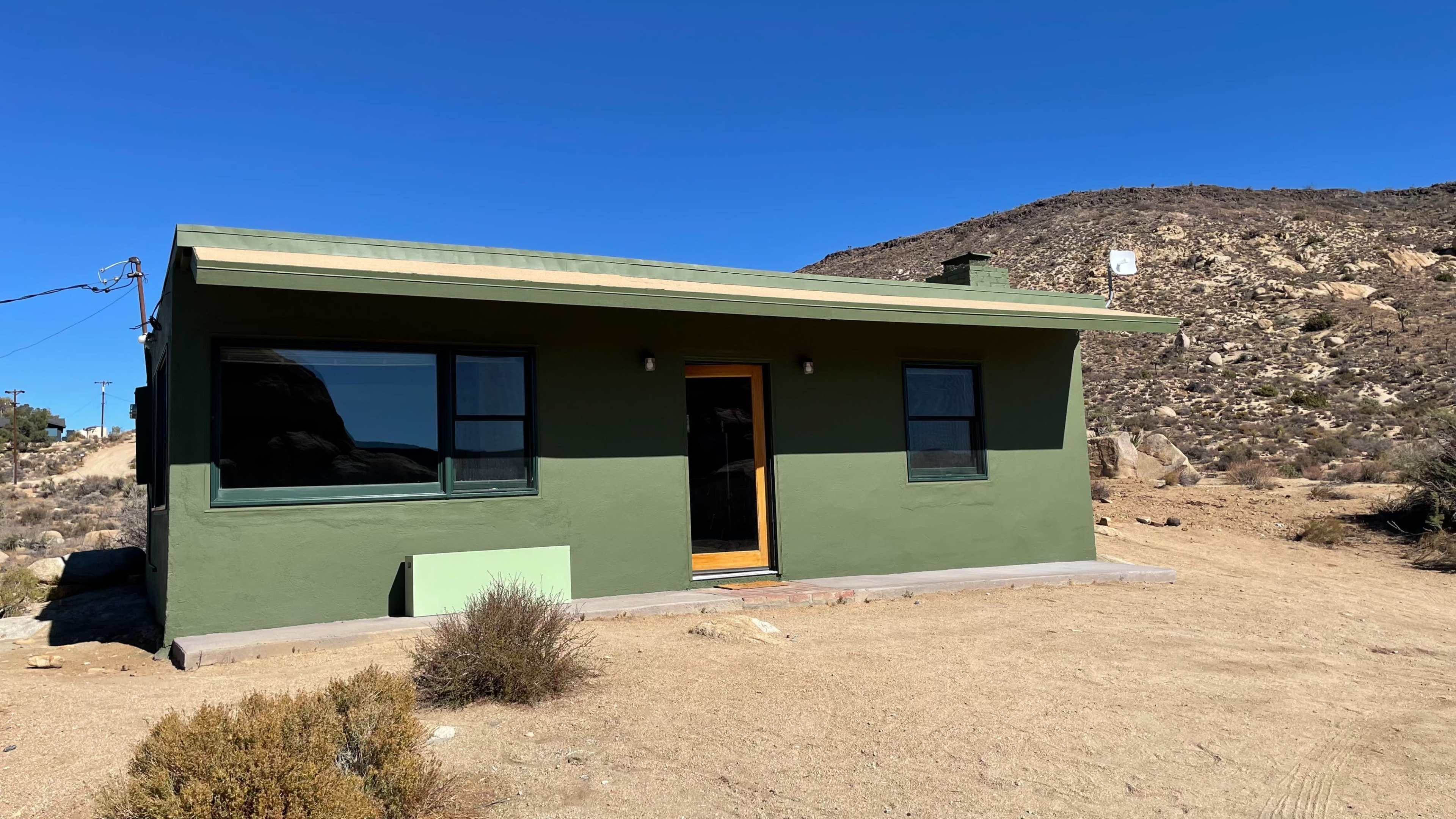 A single-story green house with large windows is situated on a sandy lot near a rocky hillside.