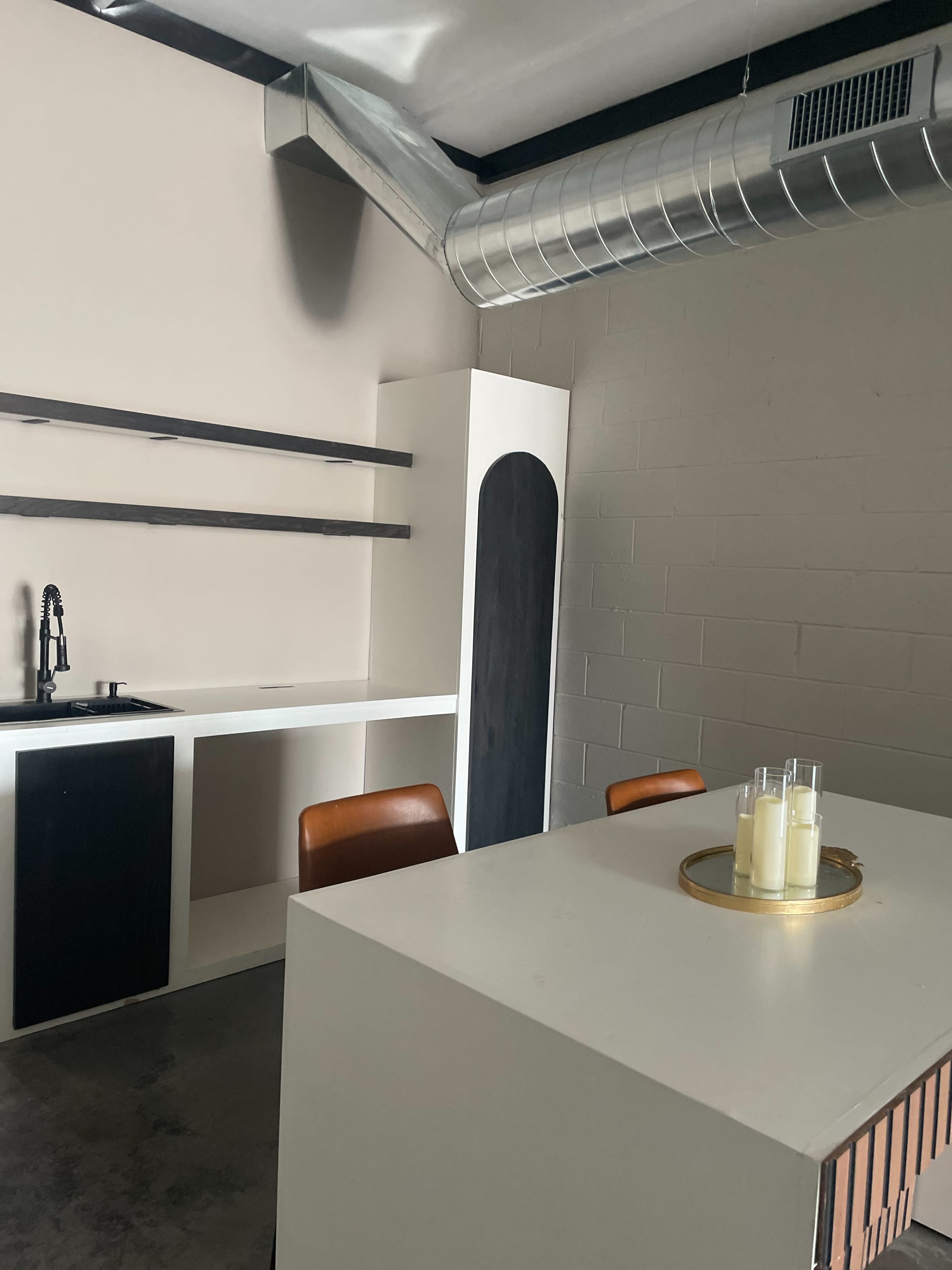 The image shows a modern, minimalist kitchen area with a white table, brown chairs, and a small set of shelves against a wall.