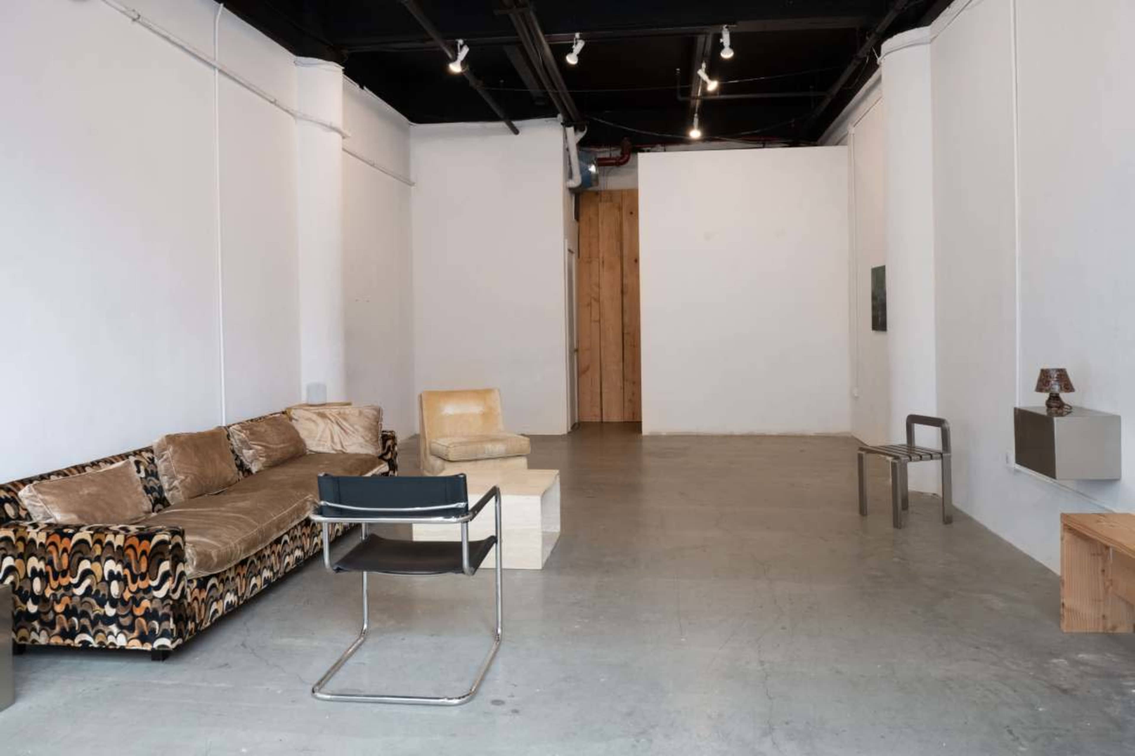 The image shows a minimalist interior space with a brown patterned sofa, a light-colored armchair, a small wooden table, and a few pieces of simple furniture.