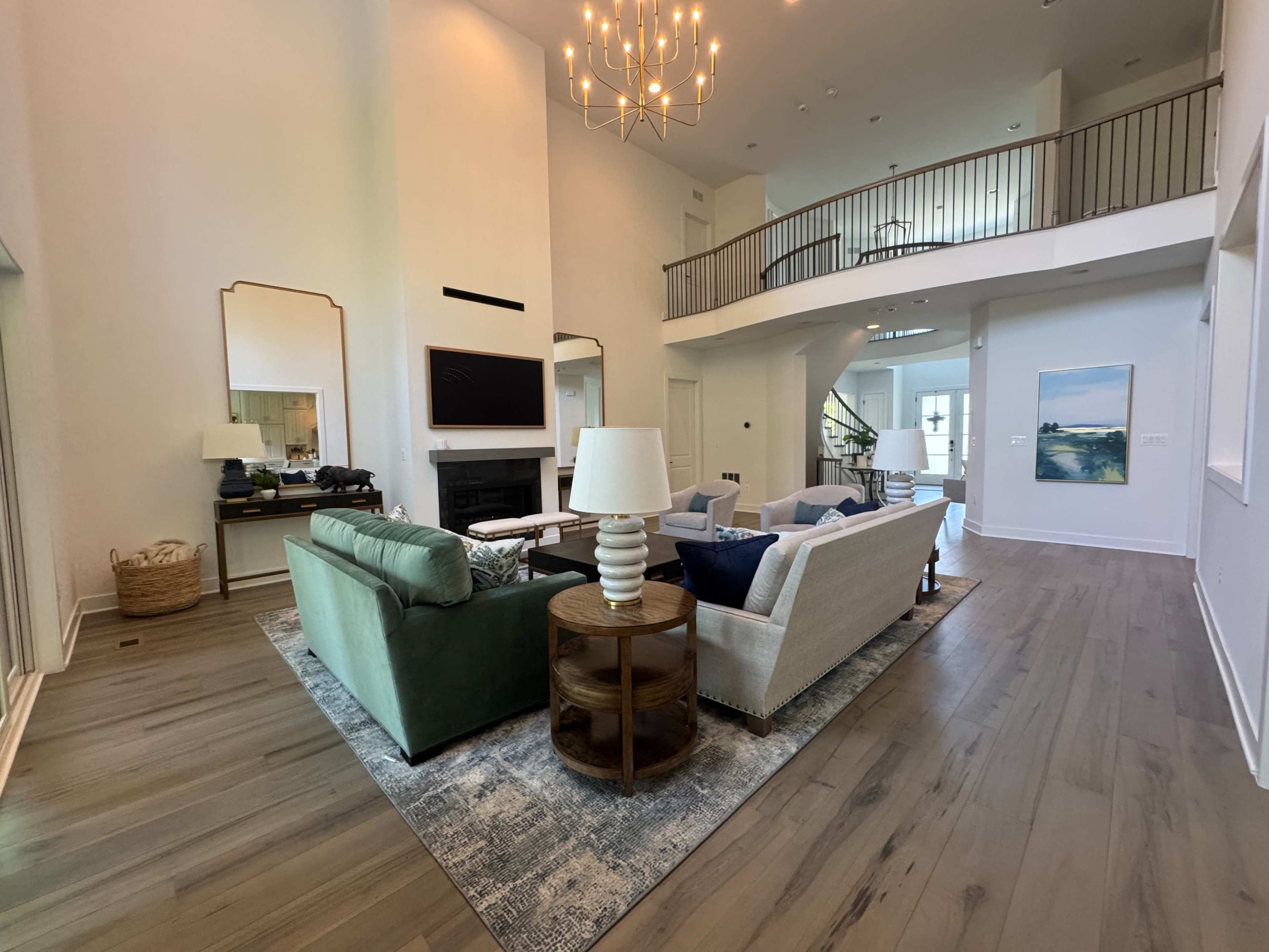 The interior of a modern living room features a seating area with a green sofa and light-colored chairs, a fireplace, and large windows that allow natural light to enter the space.
