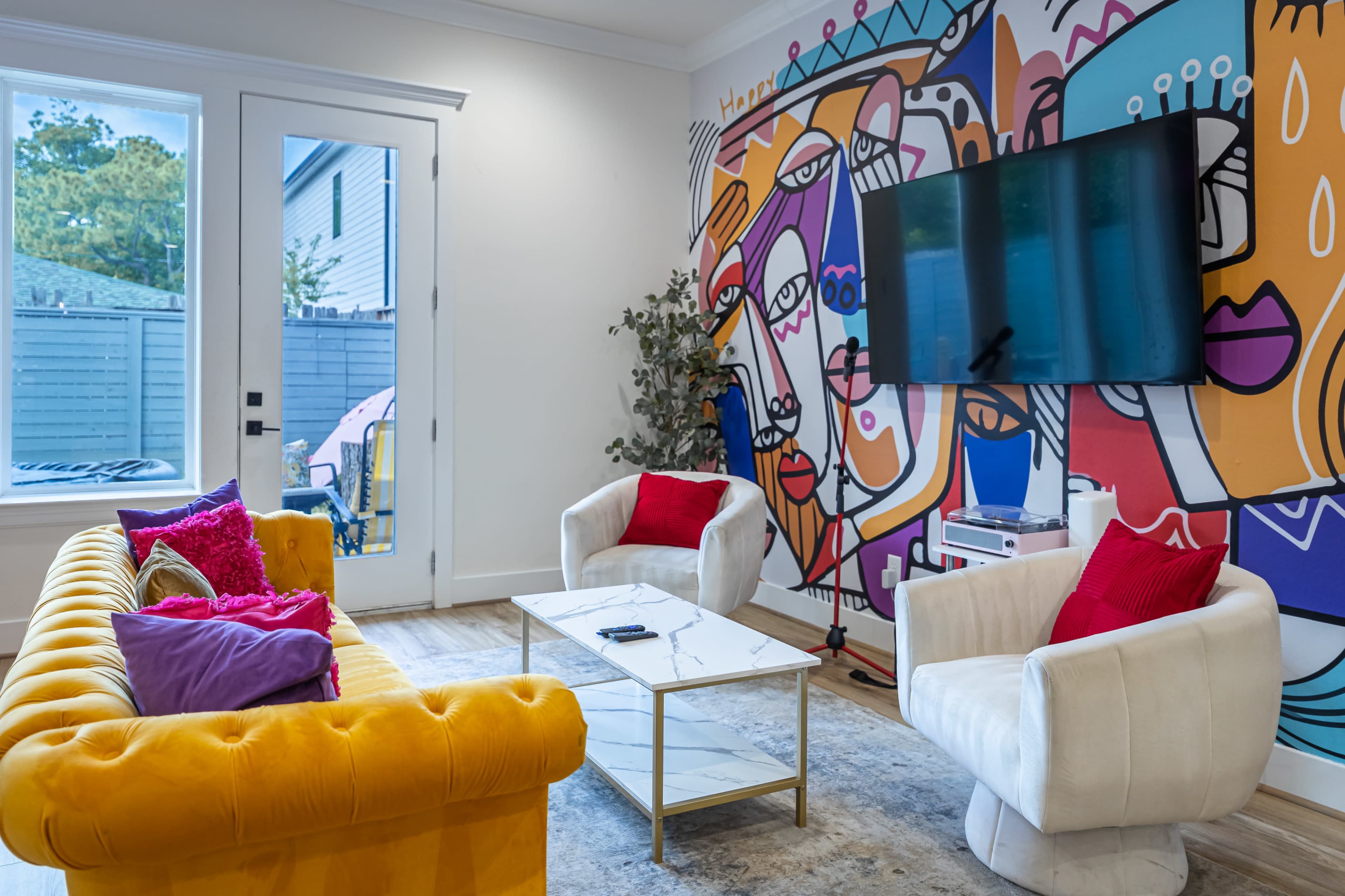The image shows a brightly decorated living room featuring a yellow sofa, two white chairs, a marble coffee table, and a colorful mural on the wall.
