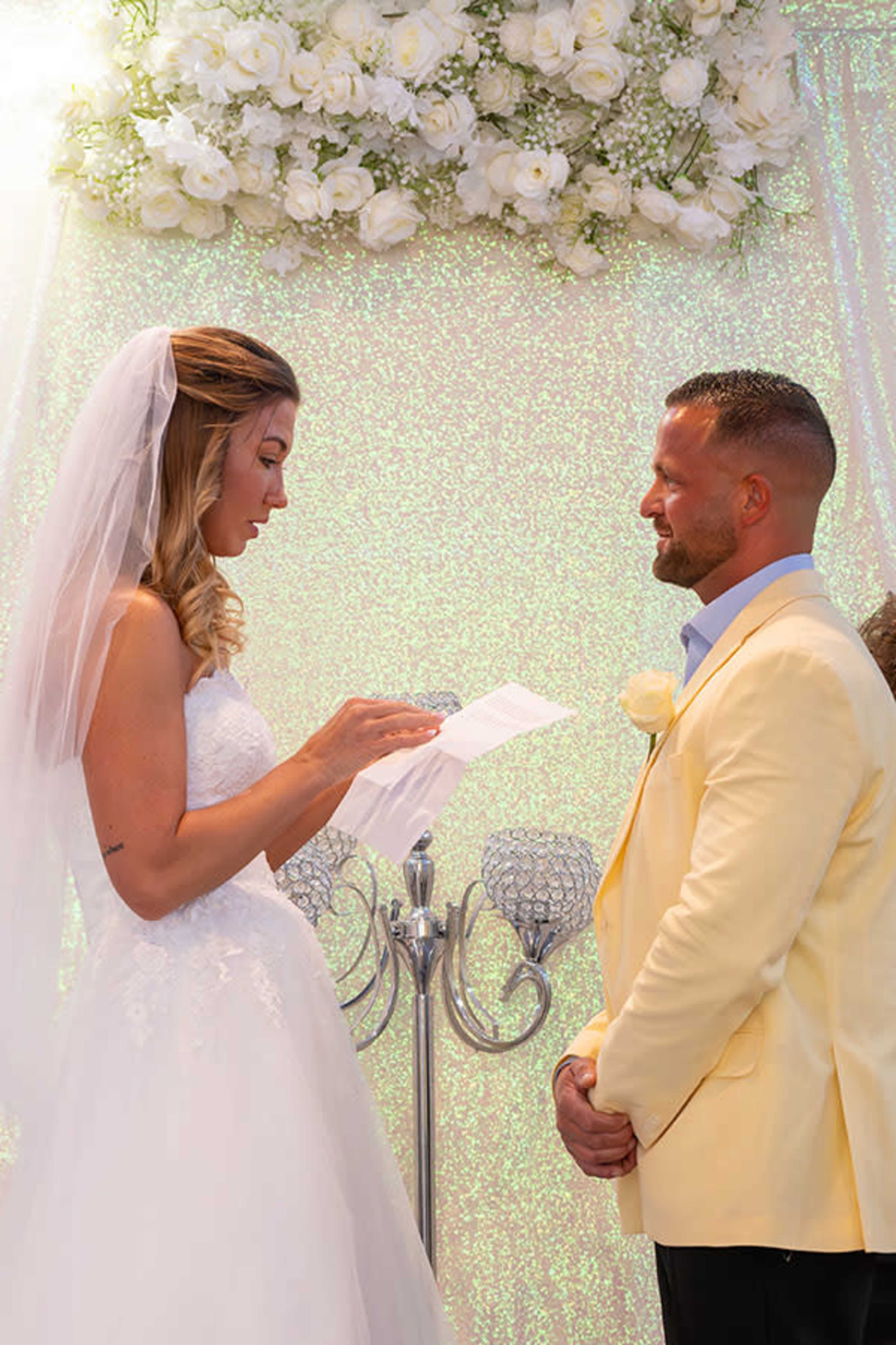 A bride in a white gown reads from a piece of paper while standing across from a man in a yellow suit during their wedding ceremony.