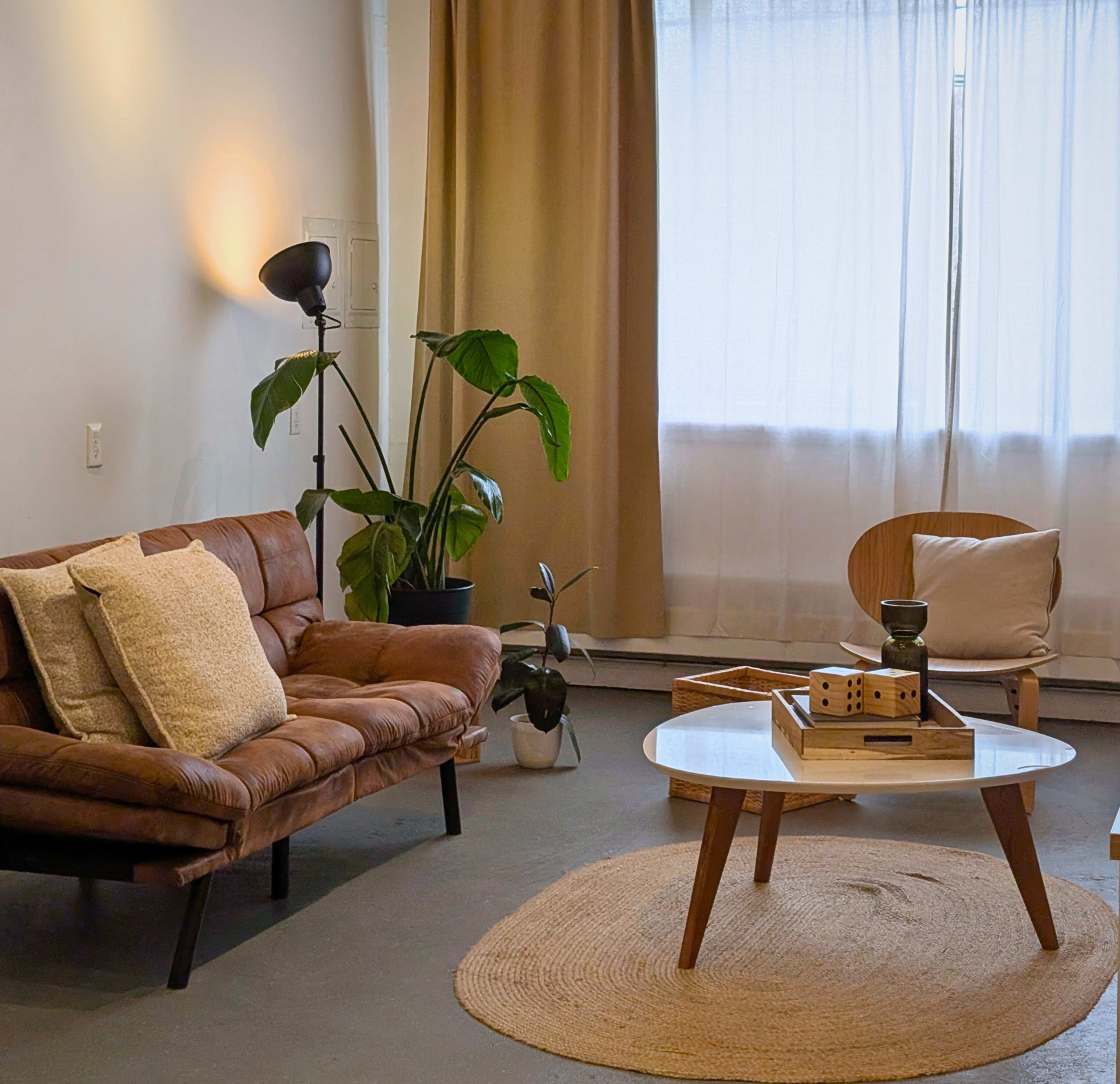 The image shows a cozy living room with a brown sofa, a round coffee table, and a large window covered with sheer curtains.