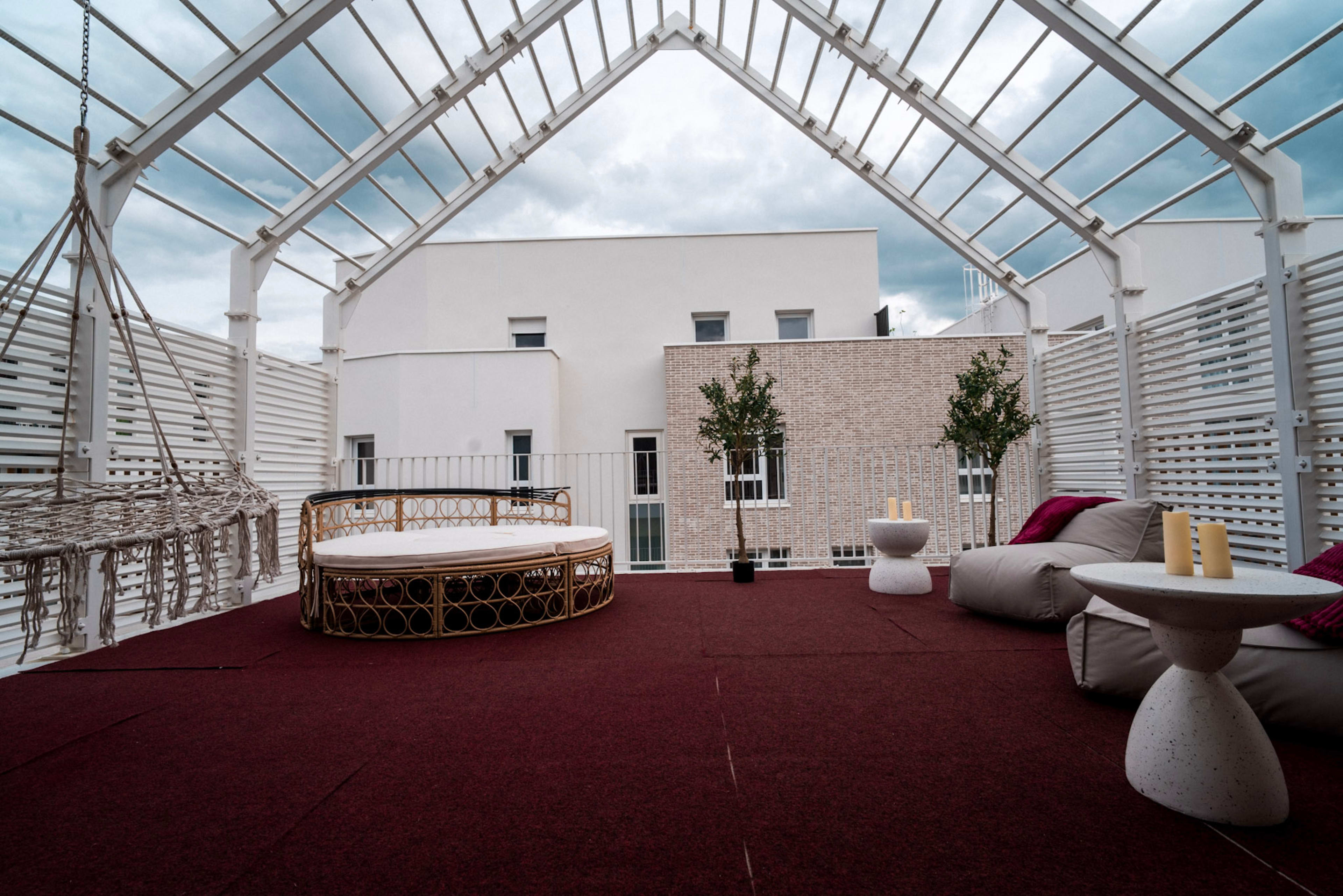 The image shows a rooftop terrace with a circular lounge bed, two side tables, a hanging chair, and potted plants, all under a transparent roof.