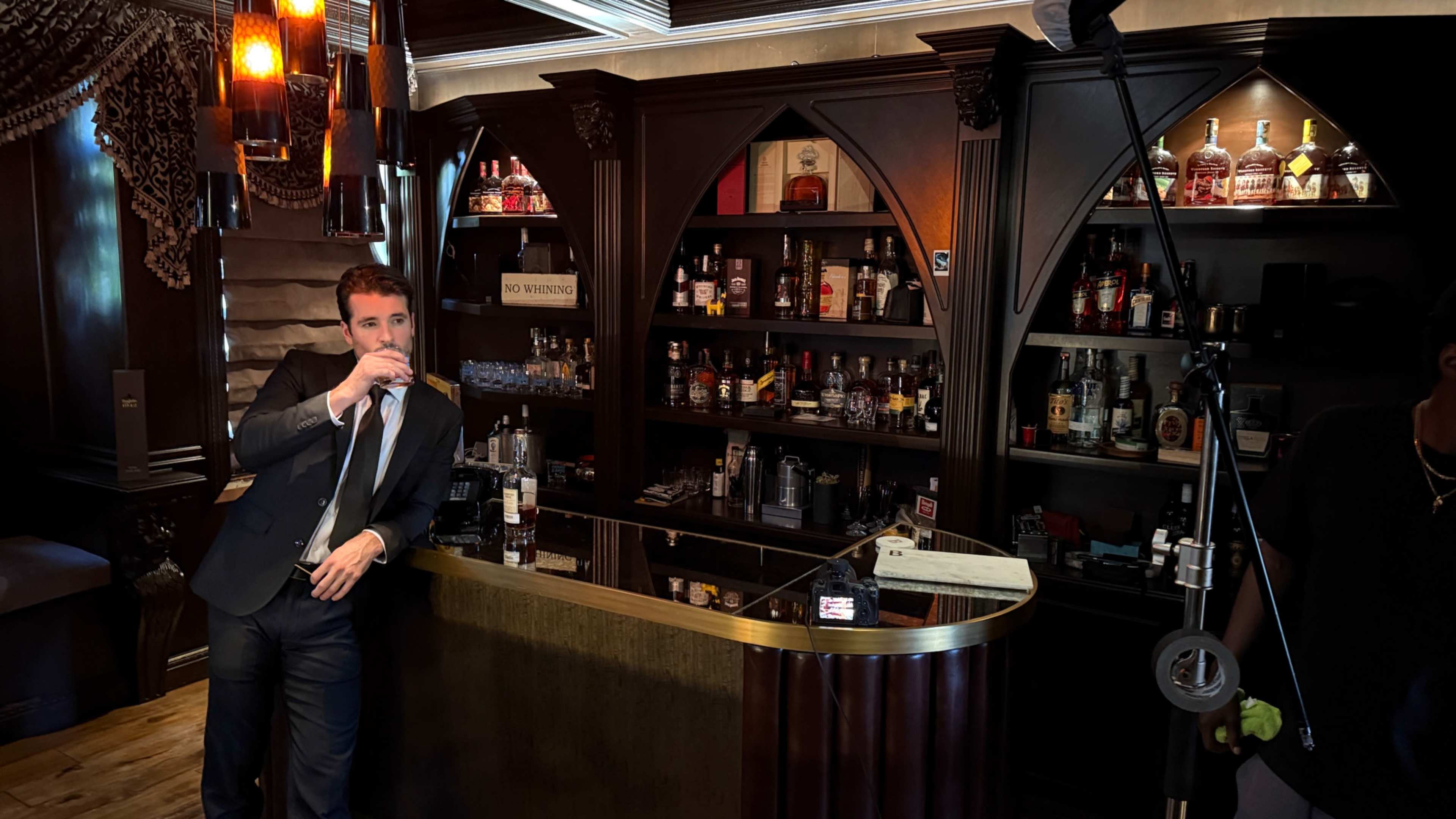 A man in a suit stands by a bar with shelves of various alcoholic beverages in a dimly lit, elegant room.