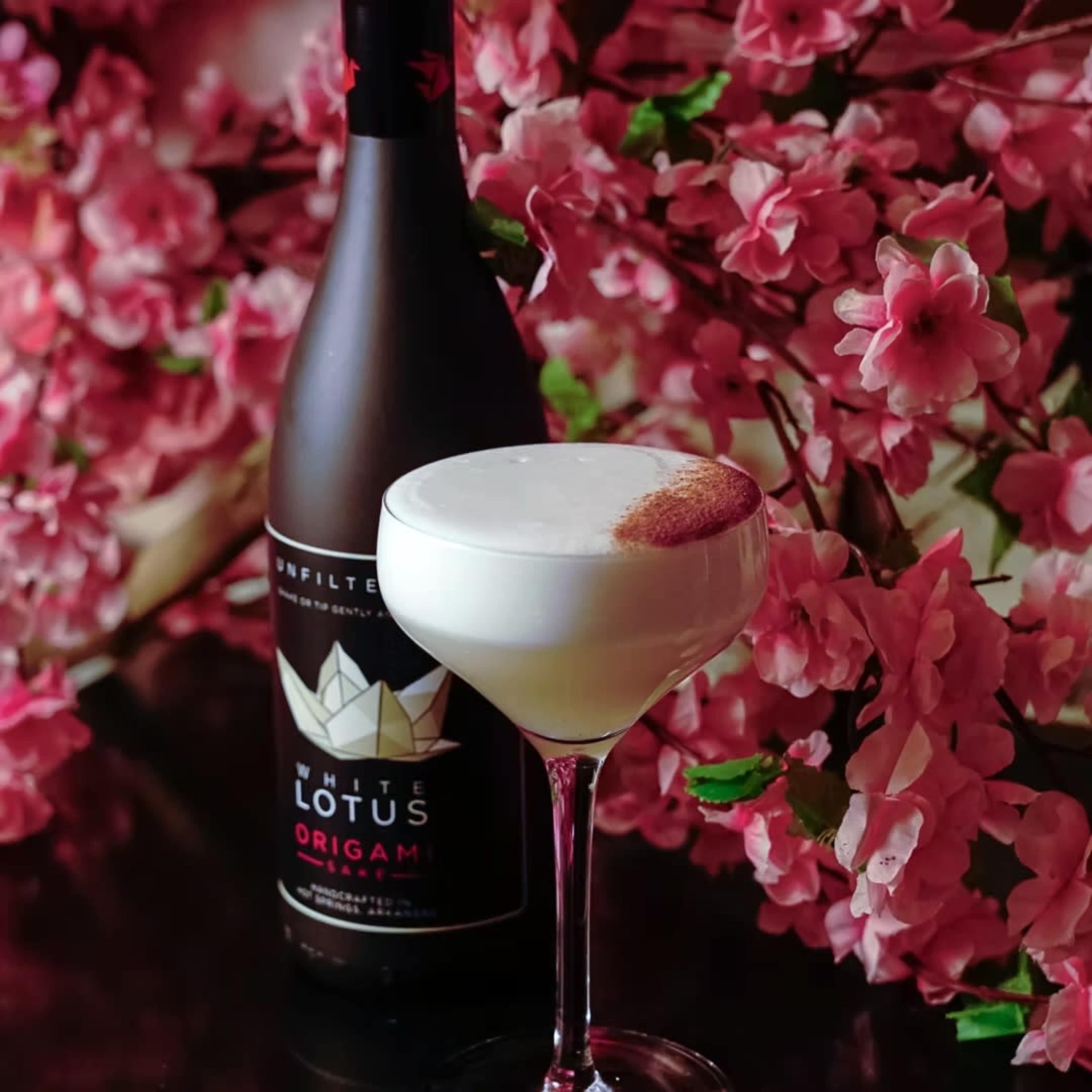 A cocktail in a stemmed glass sits beside a bottle of White Lotus Origami, with pink cherry blossoms in the background.