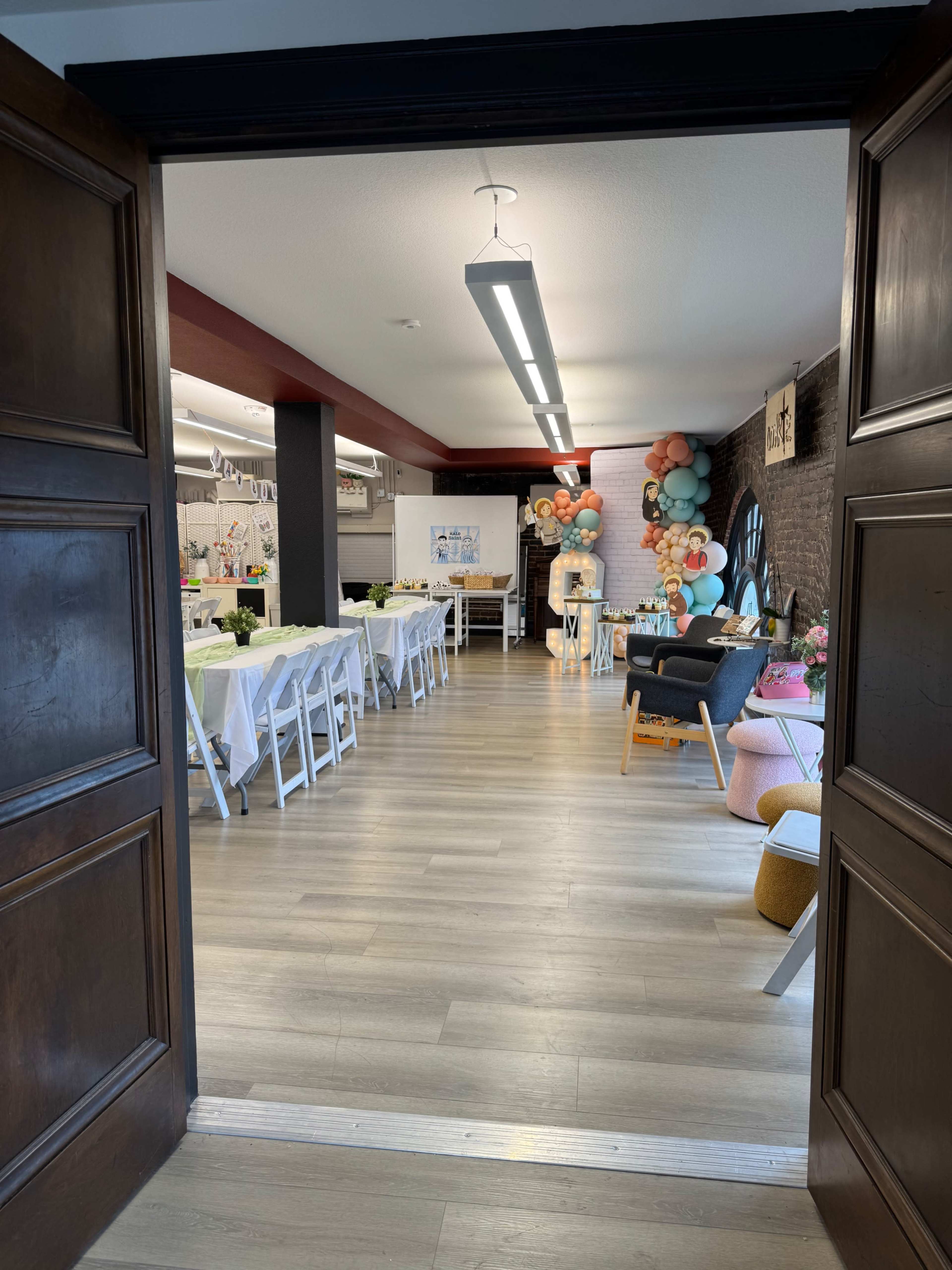 The image shows a spacious event room with tables set for a gathering, decorated with balloons and a variety of seating arrangements.