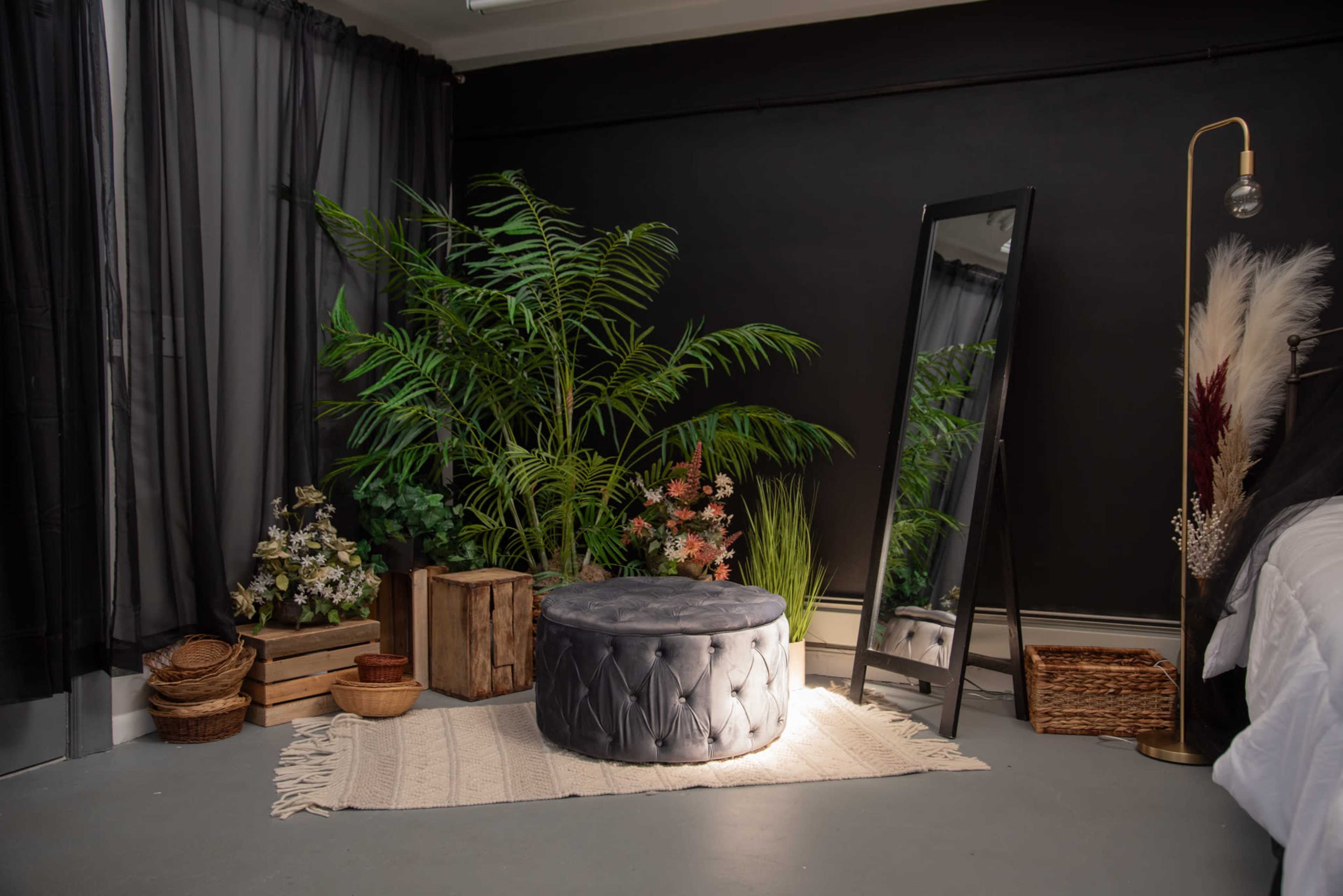 A gray tufted ottoman sits on a rug surrounded by plants, wooden crates, and a tall mirror in a dimly lit room.