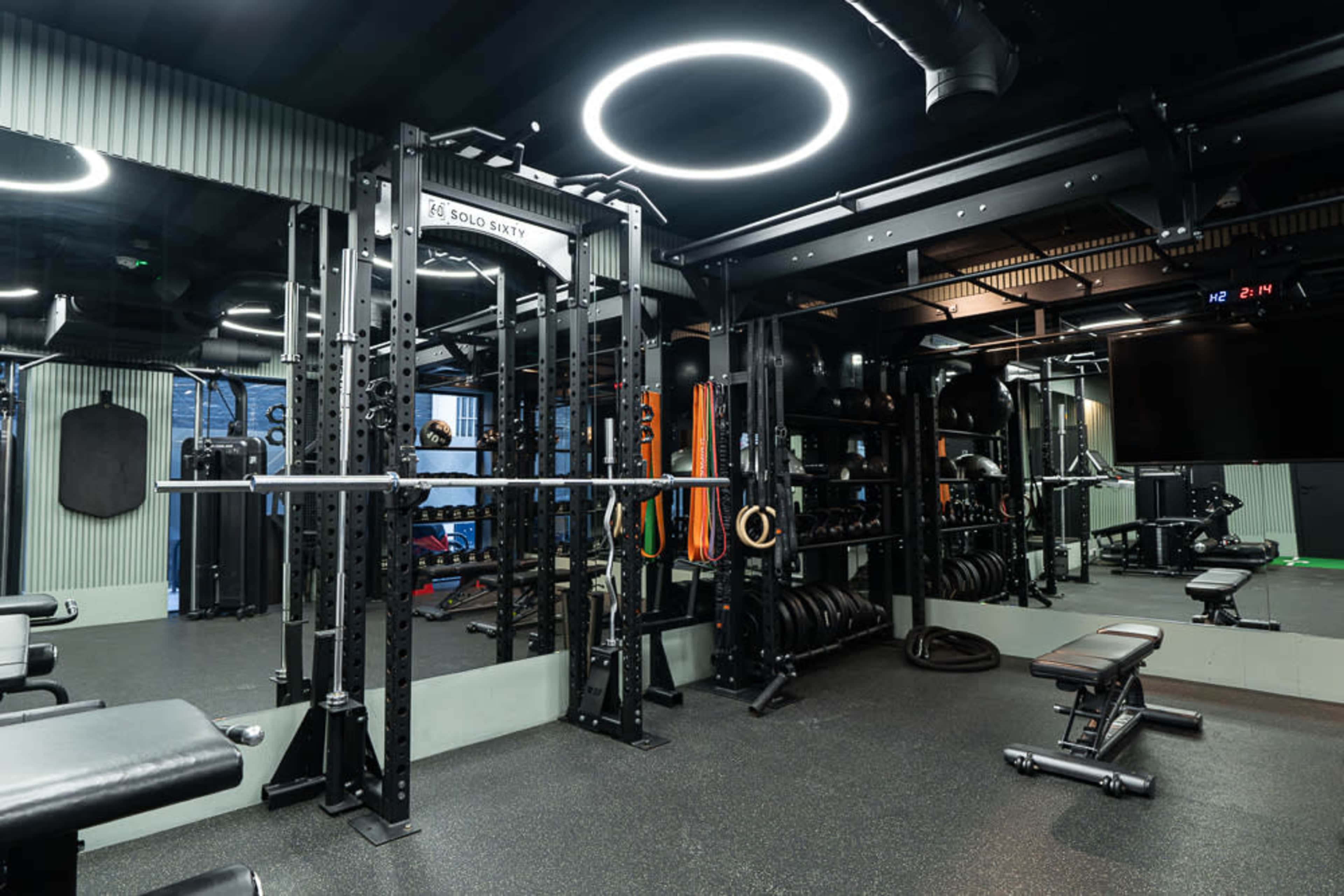 The image shows a modern gym equipped with various fitness equipment, including weightlifting racks, benches, and a collection of free weights.