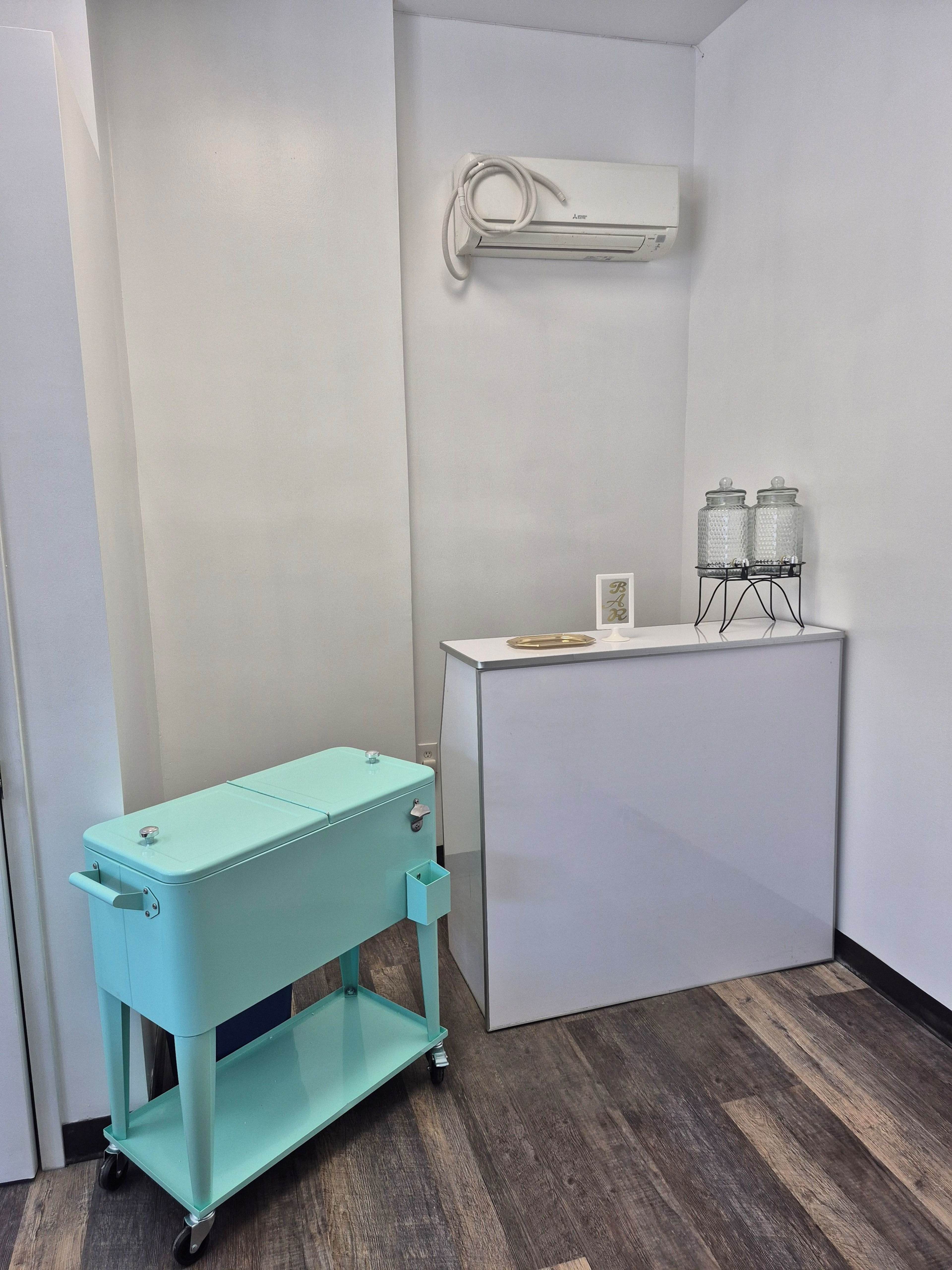 The image shows a small, light blue cart on wheels next to a white cabinet and decorative glass containers against a plain wall.