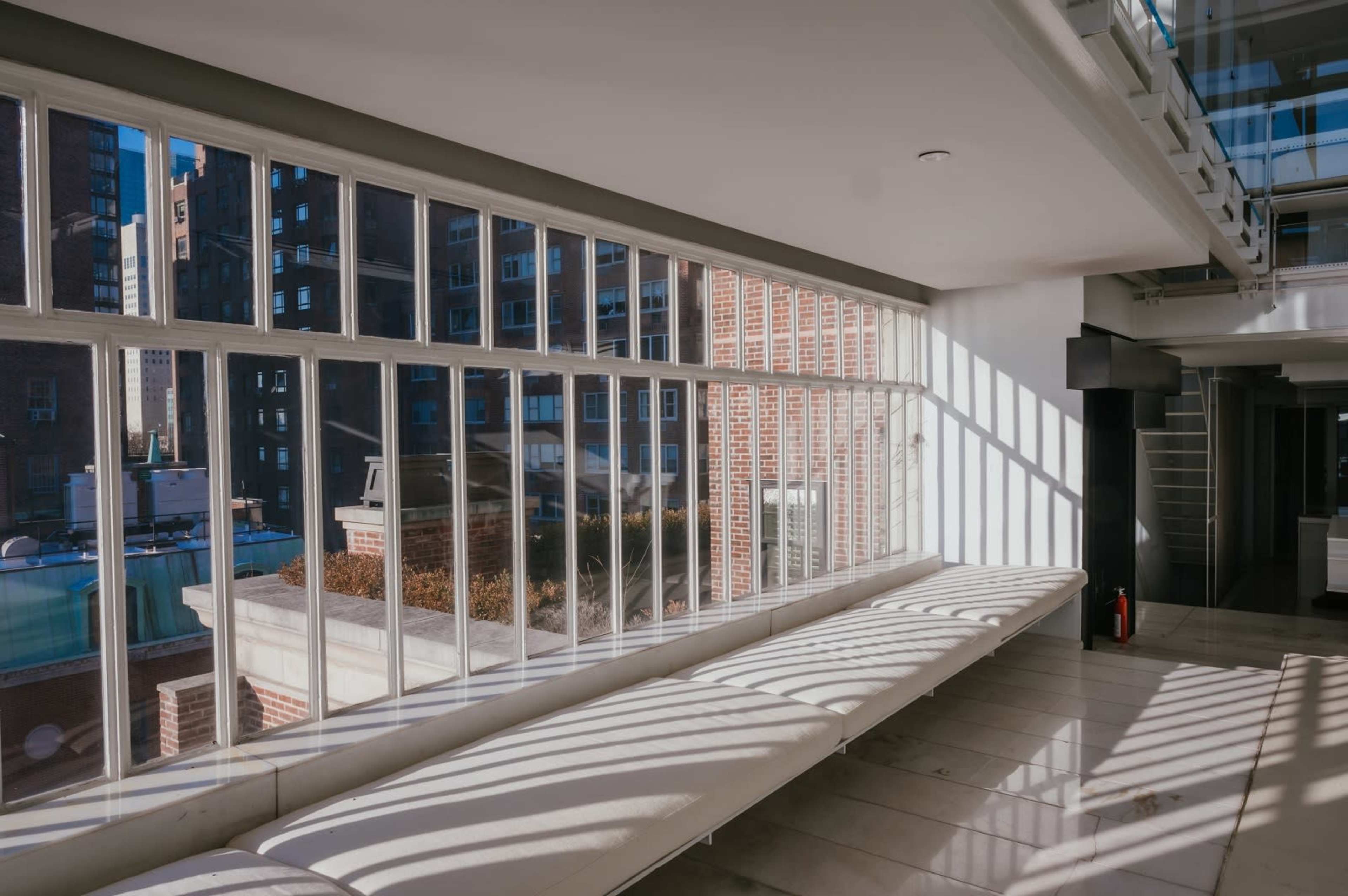 The image shows a modern interior space with large windows allowing natural light to cast shadows on a long, white bench.