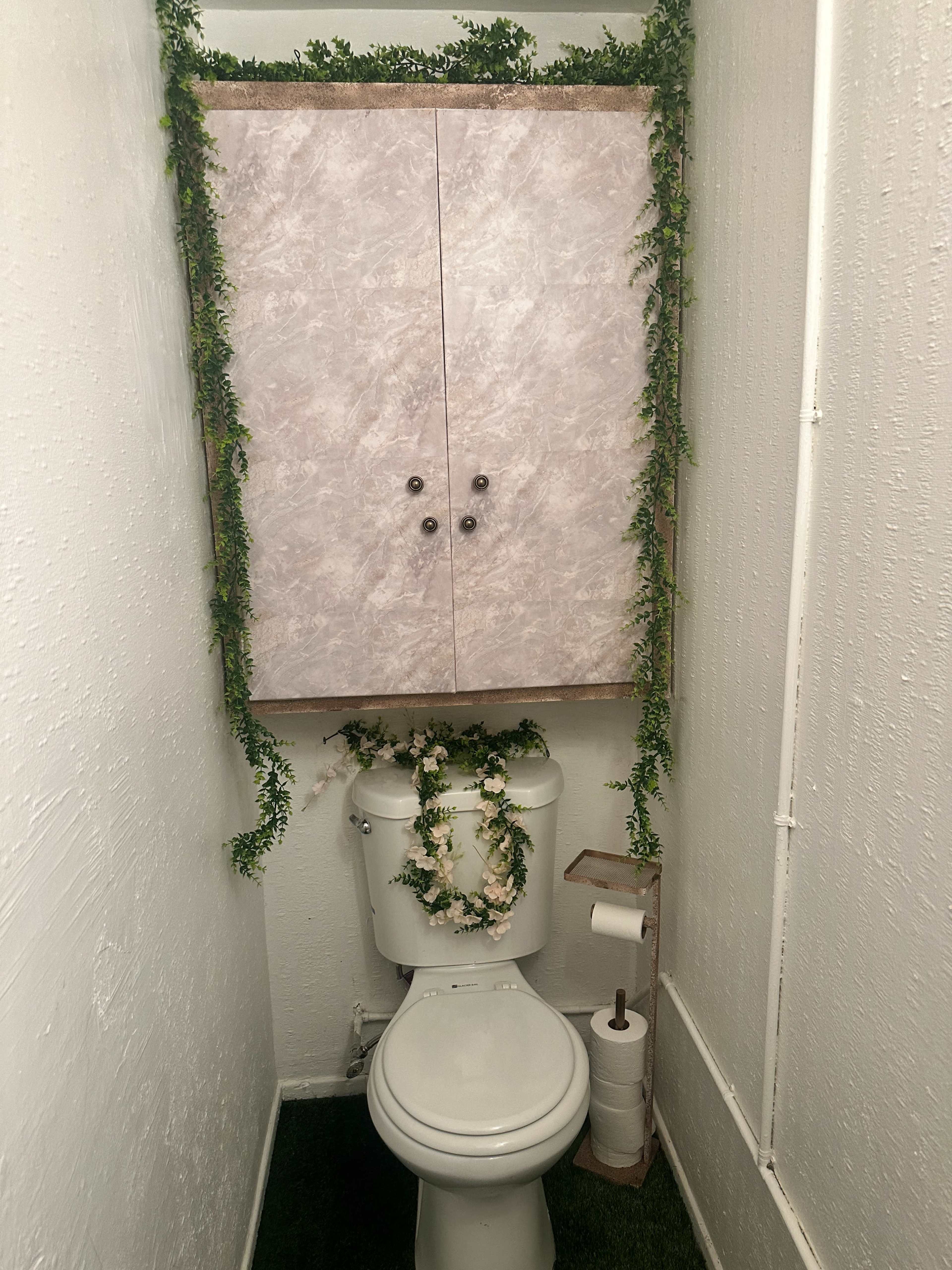 A small bathroom with a toilet beneath a cabinet decorated with faux greenery.
