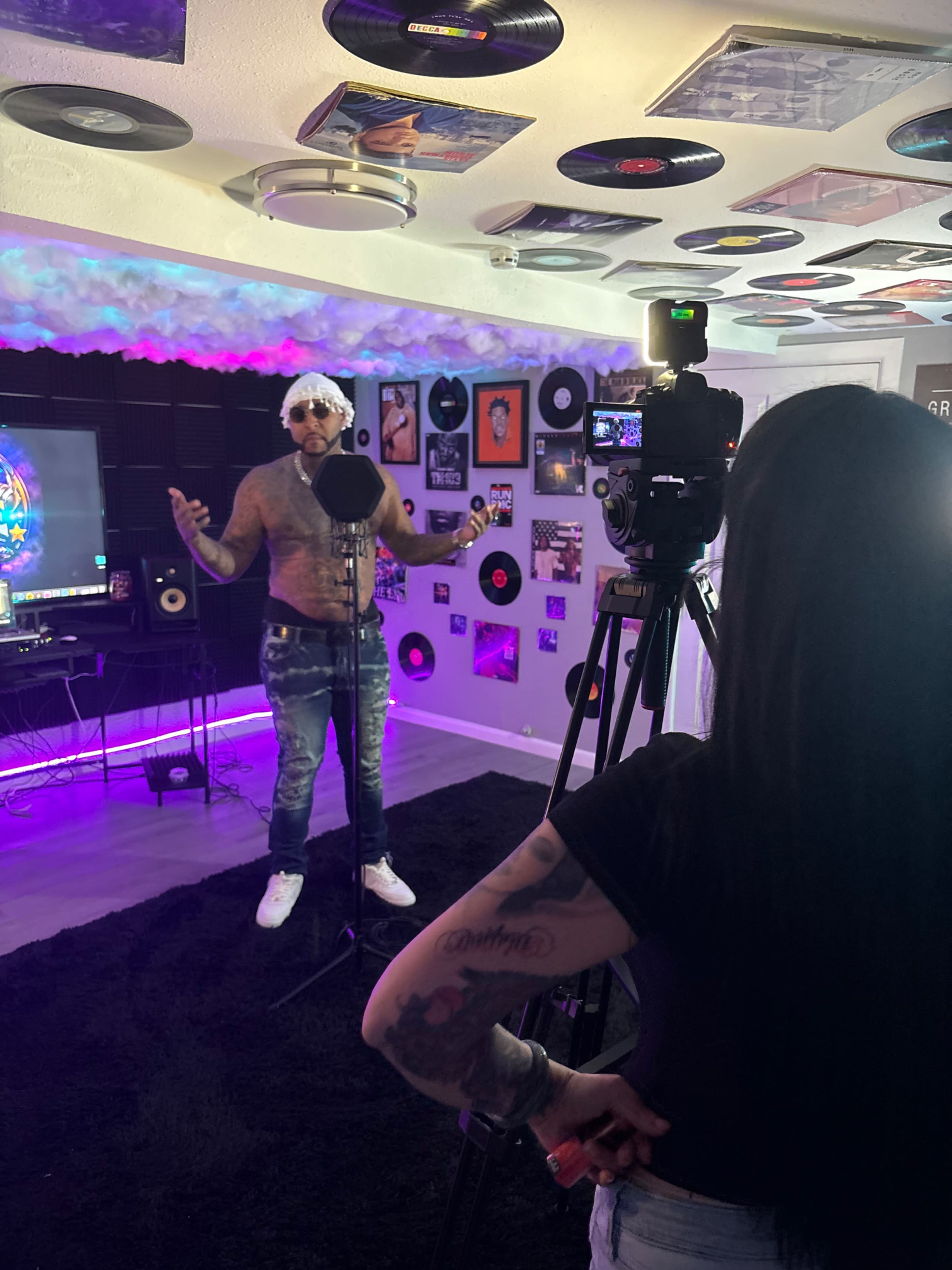 A man with a styled appearance is performing in front of a camera while a woman records him in a room decorated with vinyl records and colorful lighting.