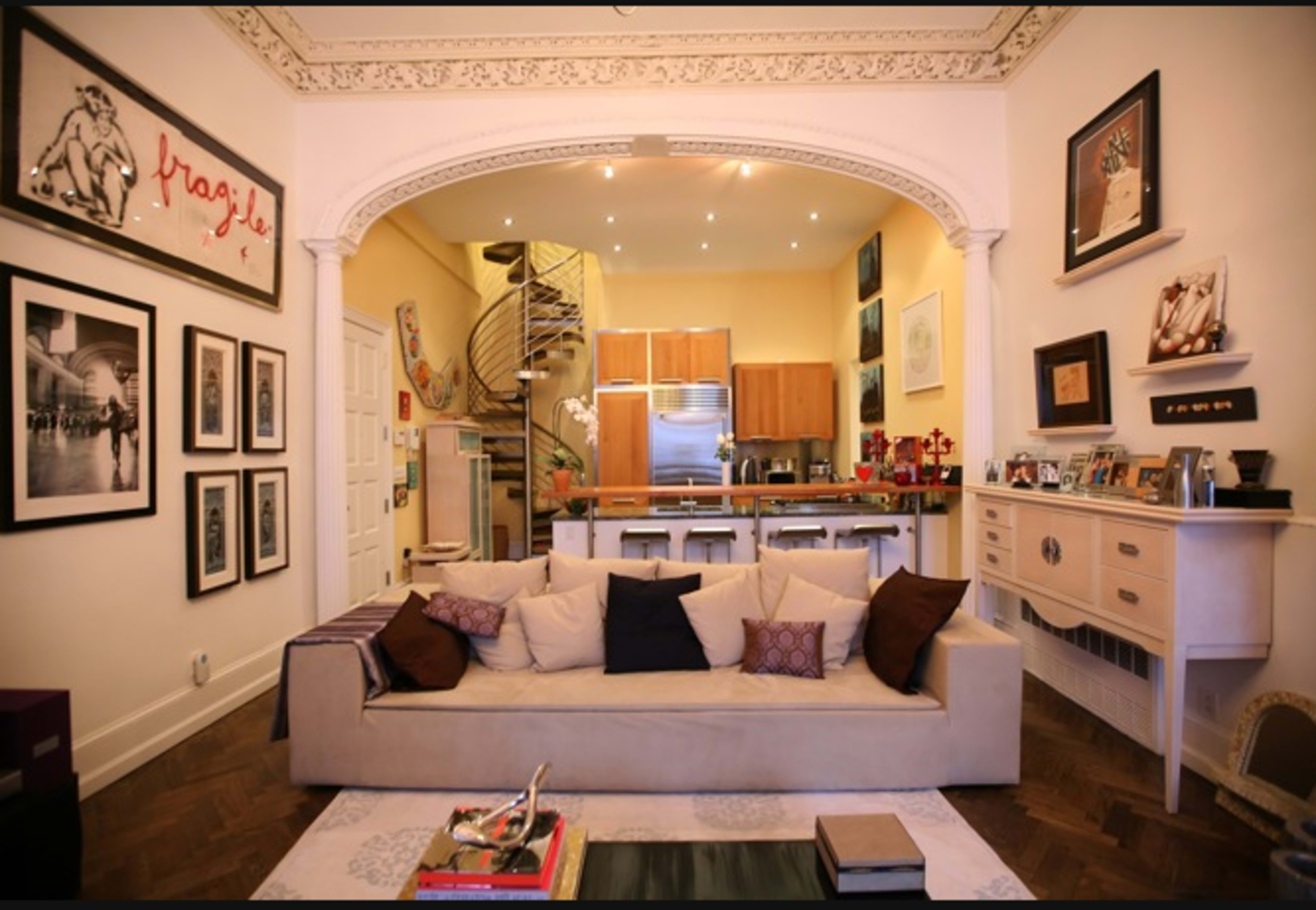 The image shows a well-decorated living room with a large beige sofa, framed photographs on the walls, and a view of a modern kitchen in the background.