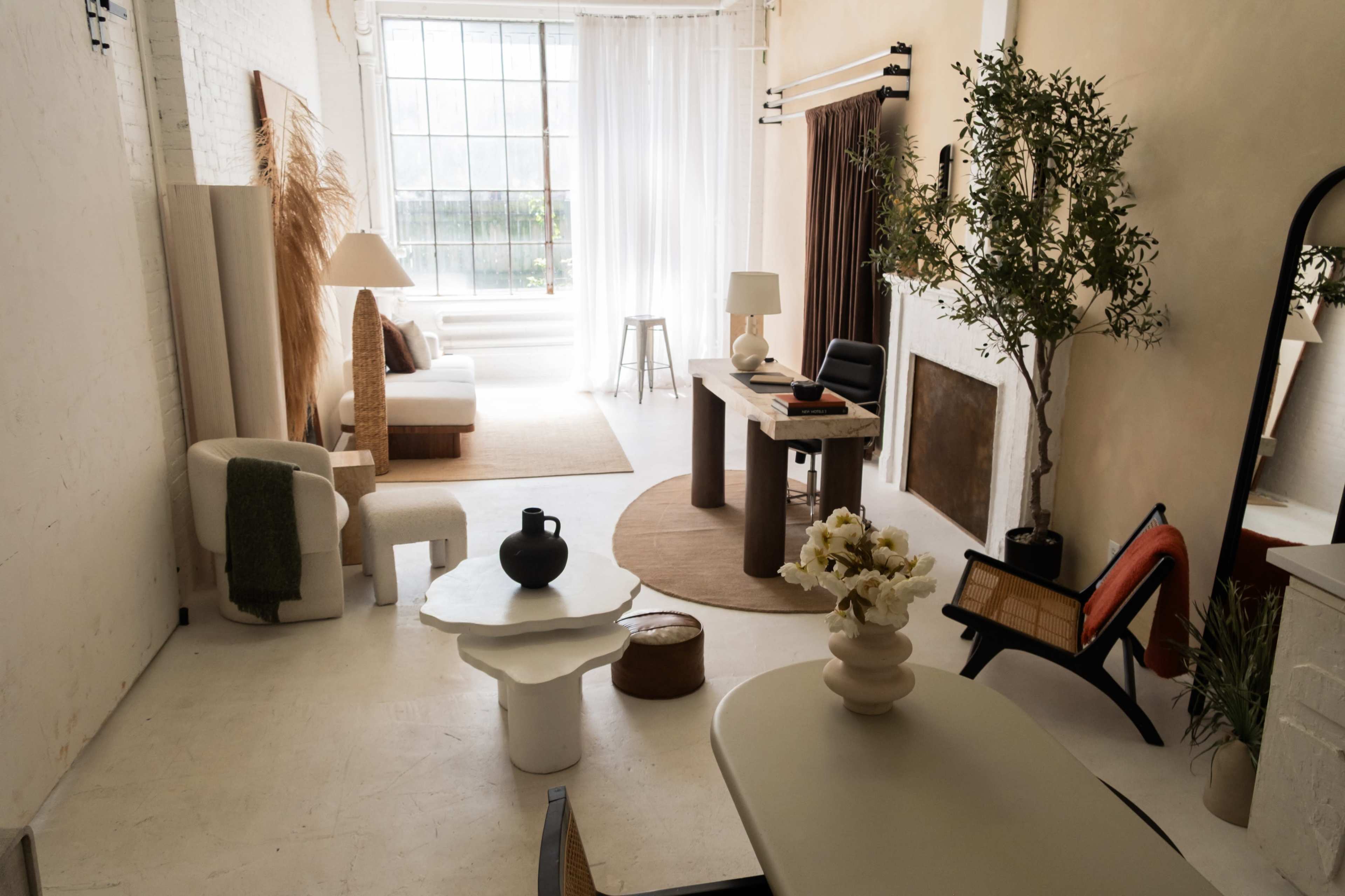 The image shows a minimalist interior space featuring a living area with a sofa, a desk, various decorative elements, and large windows that allow natural light to enter.