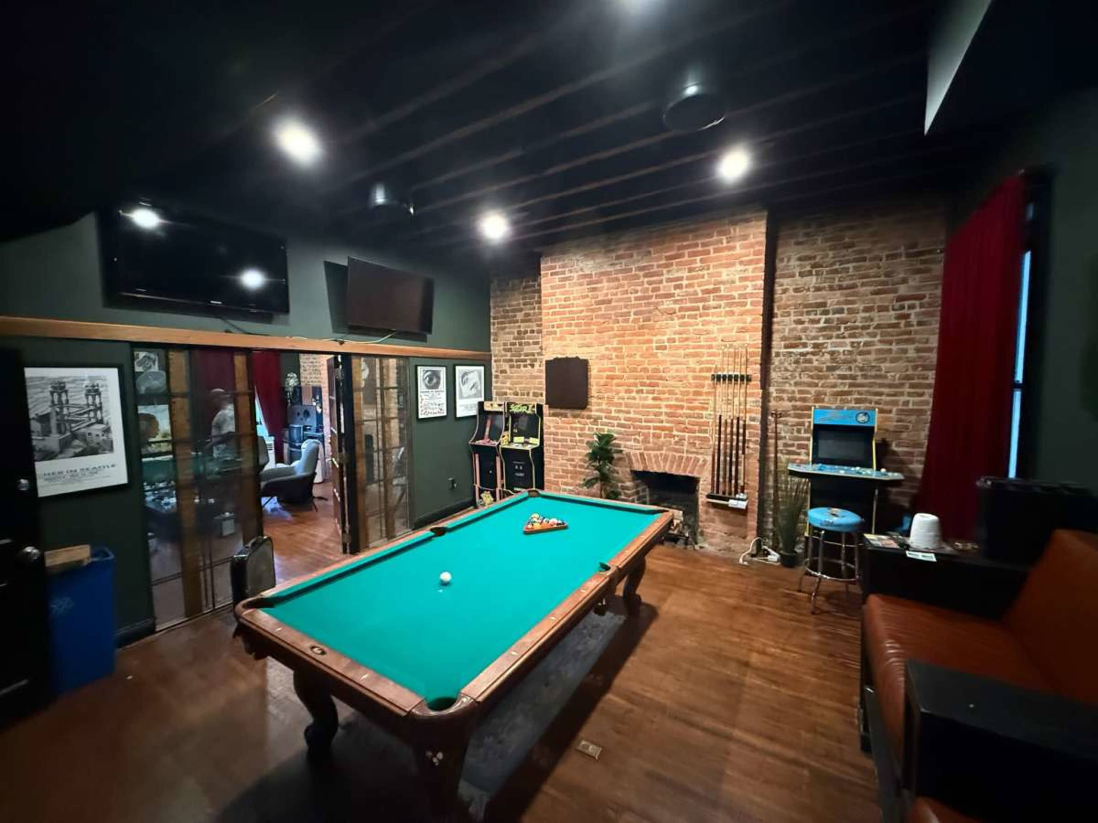 The image shows a room with a green pool table in the center, brick walls, and various recreational equipment along with a seating area.