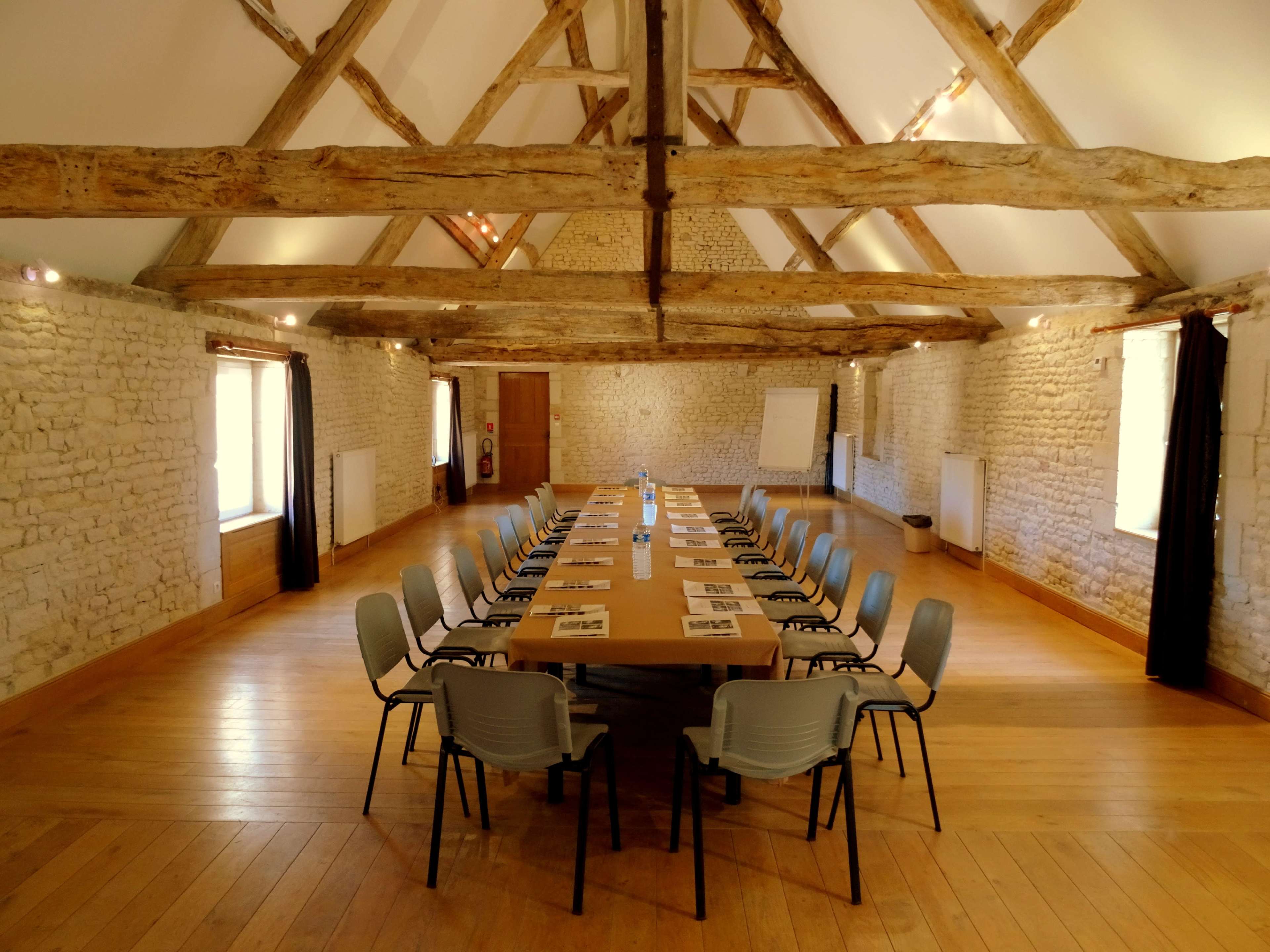A long wooden table is surrounded by gray chairs in a spacious room with exposed wooden beams and stone walls.