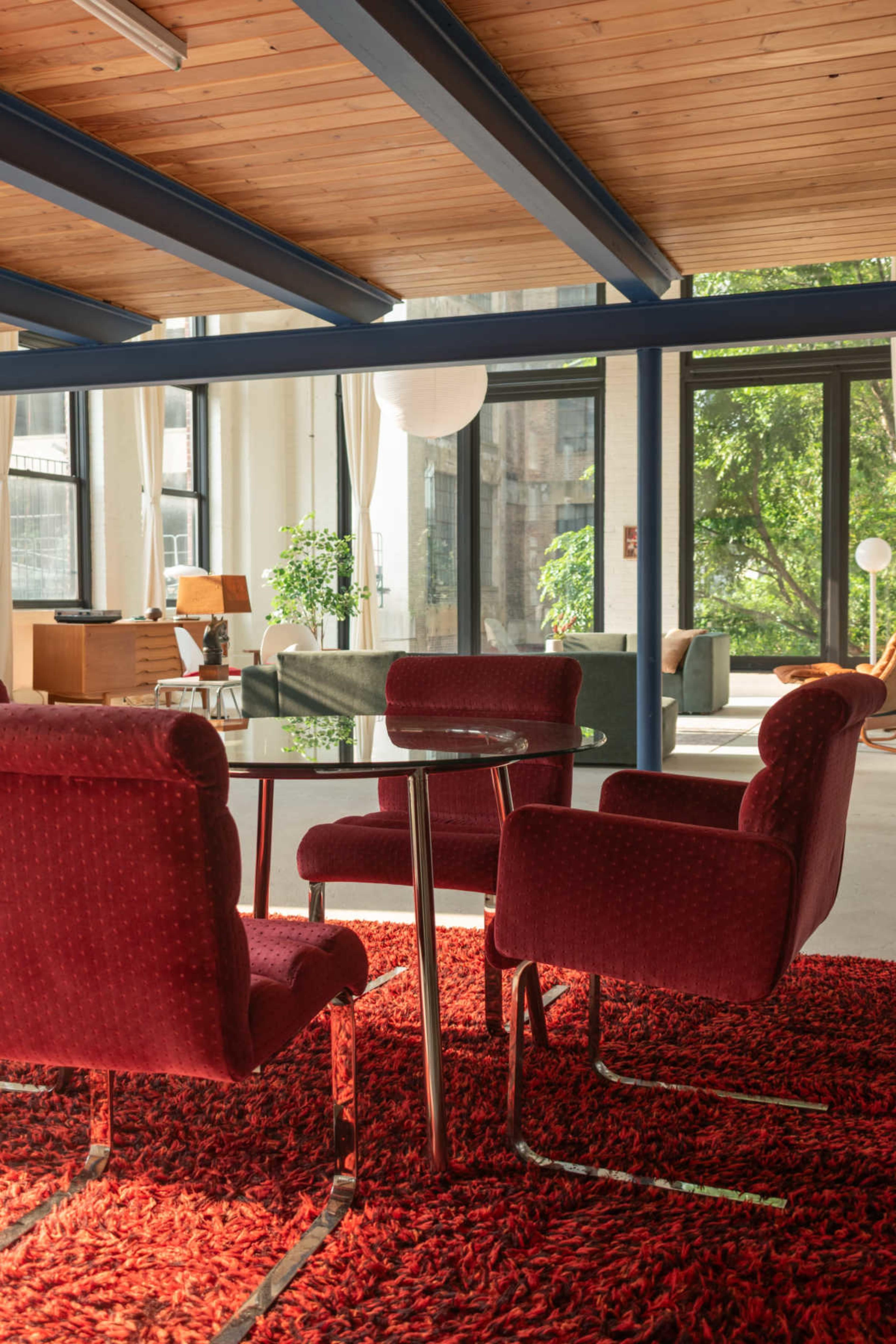 A bright room features a glass table surrounded by four red upholstered chairs on a textured red rug, with large windows allowing natural light to illuminate the space.