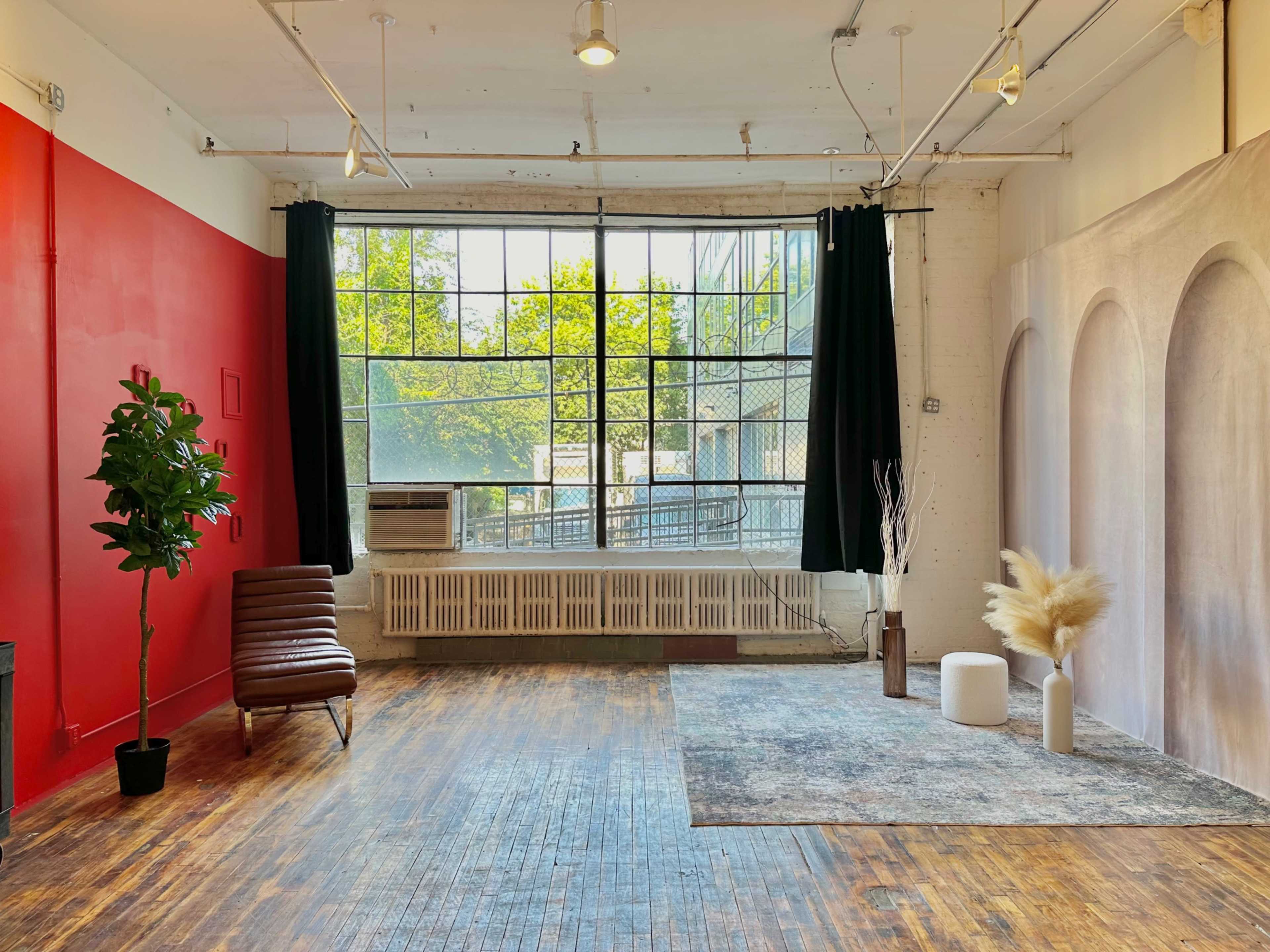 The image showcases a spacious room with large windows, wooden flooring, and minimalist decor elements, including a green plant, a leather chair, and decorative vases.