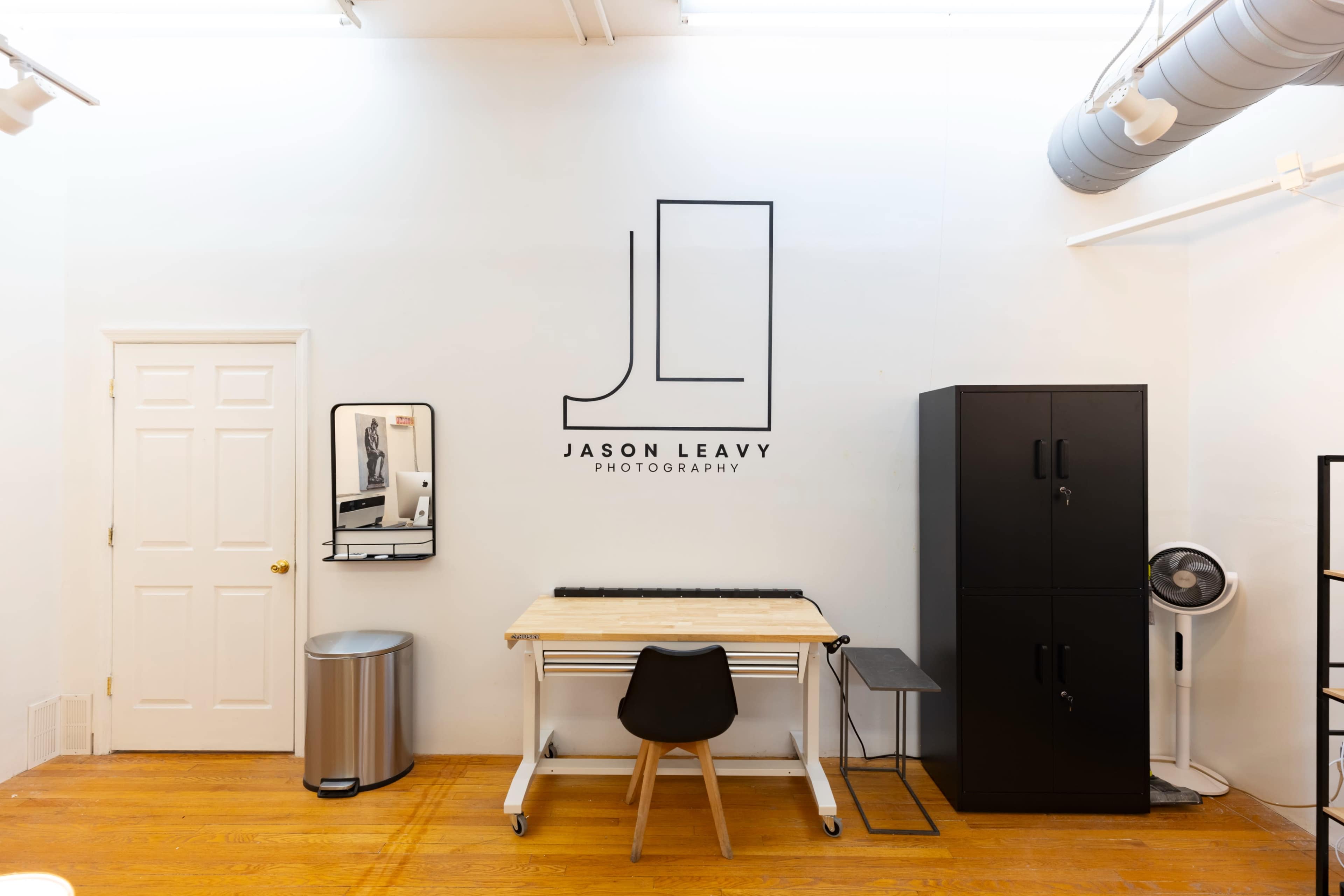 The image shows a clean photography studio with a central work table, a black cabinet, a mirror, and a door along the wall featuring the logo "JASON LEAVY PHOTOGRAPHY."