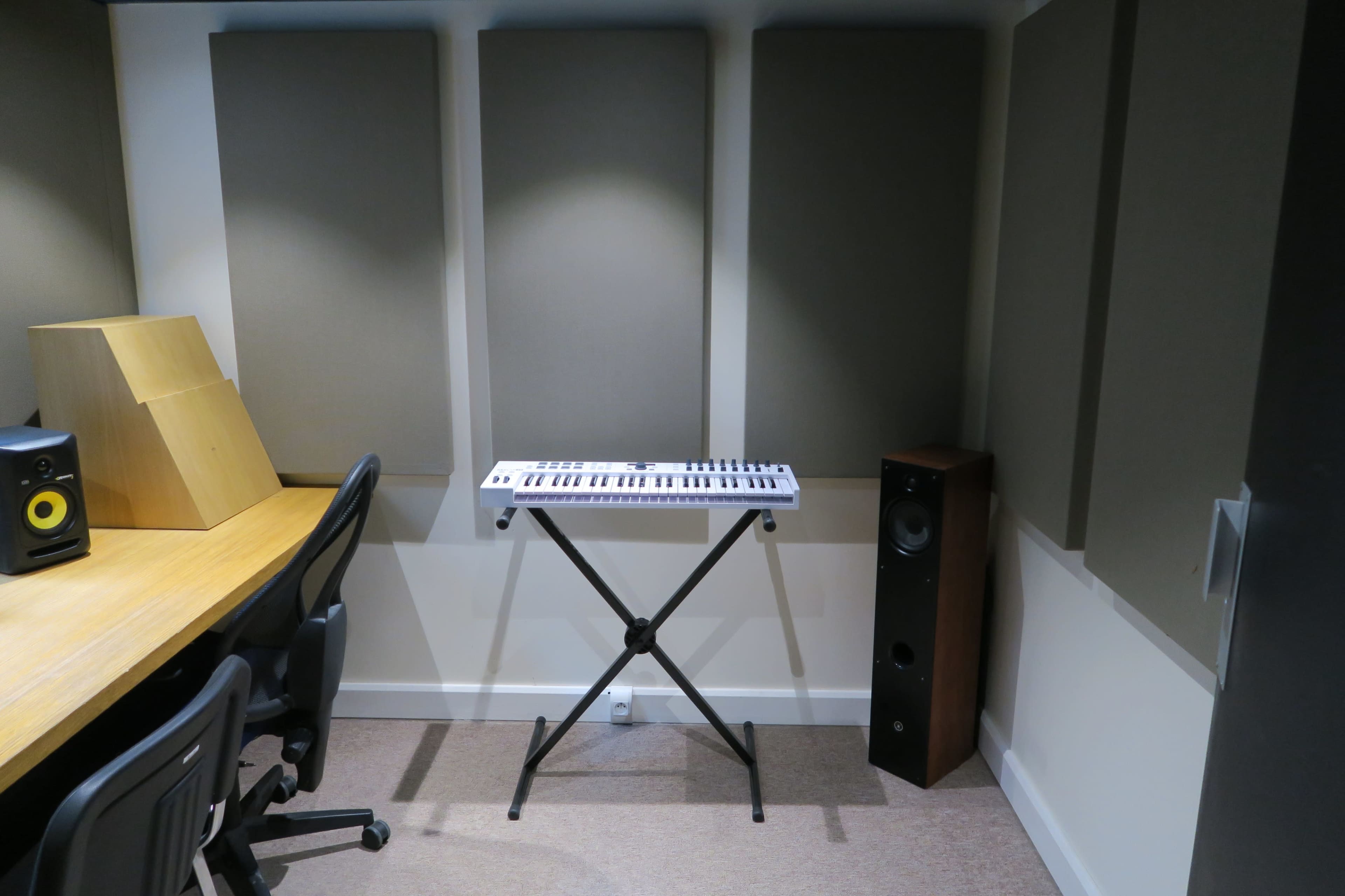 The image shows a small music studio with a keyboard on a stand, surrounded by soundproof wall panels, a wooden desk, and a speaker in the corner.