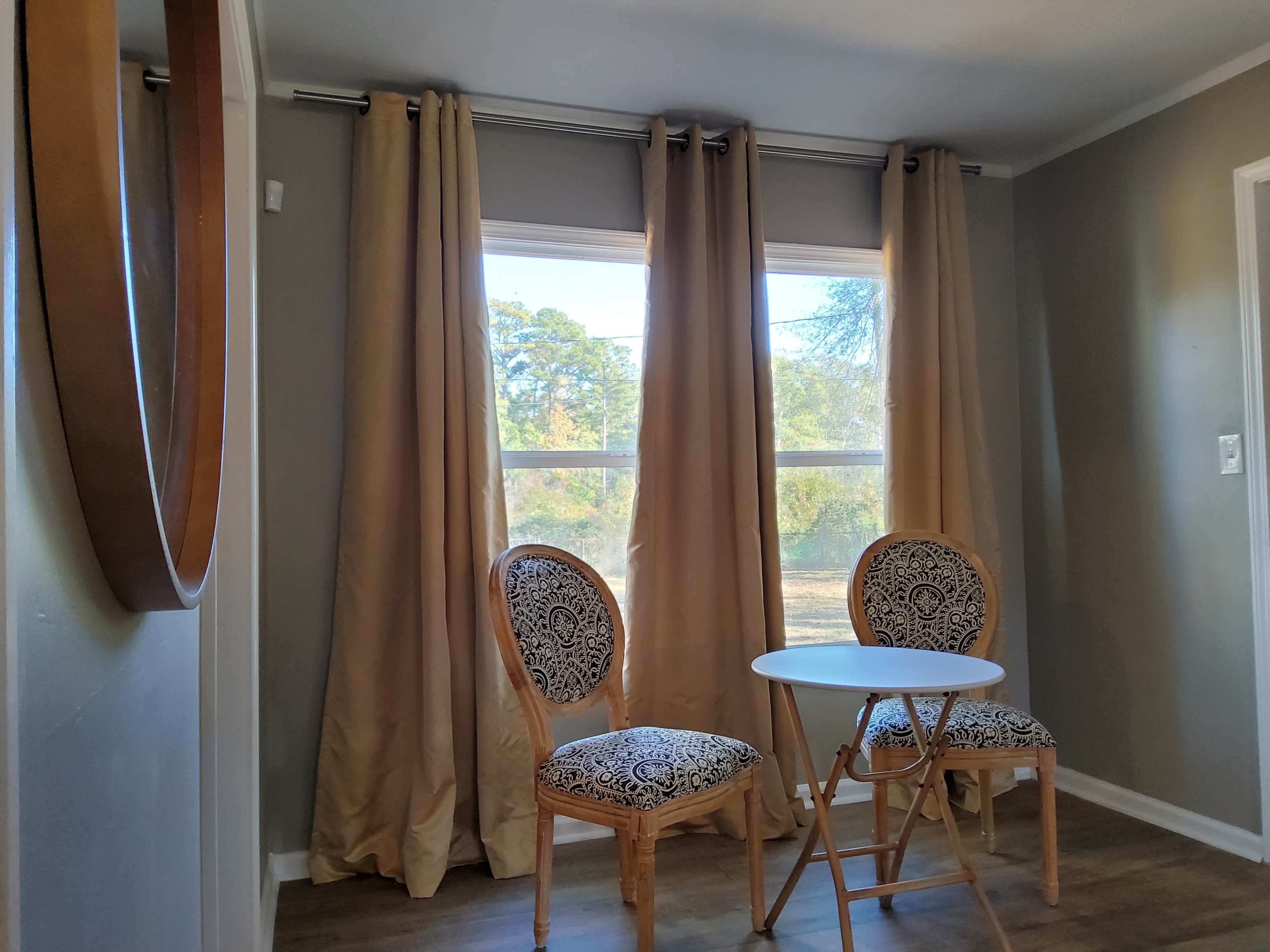 A small seating area with two patterned chairs positioned around a white circular table, set against a backdrop of large windows with beige curtains.