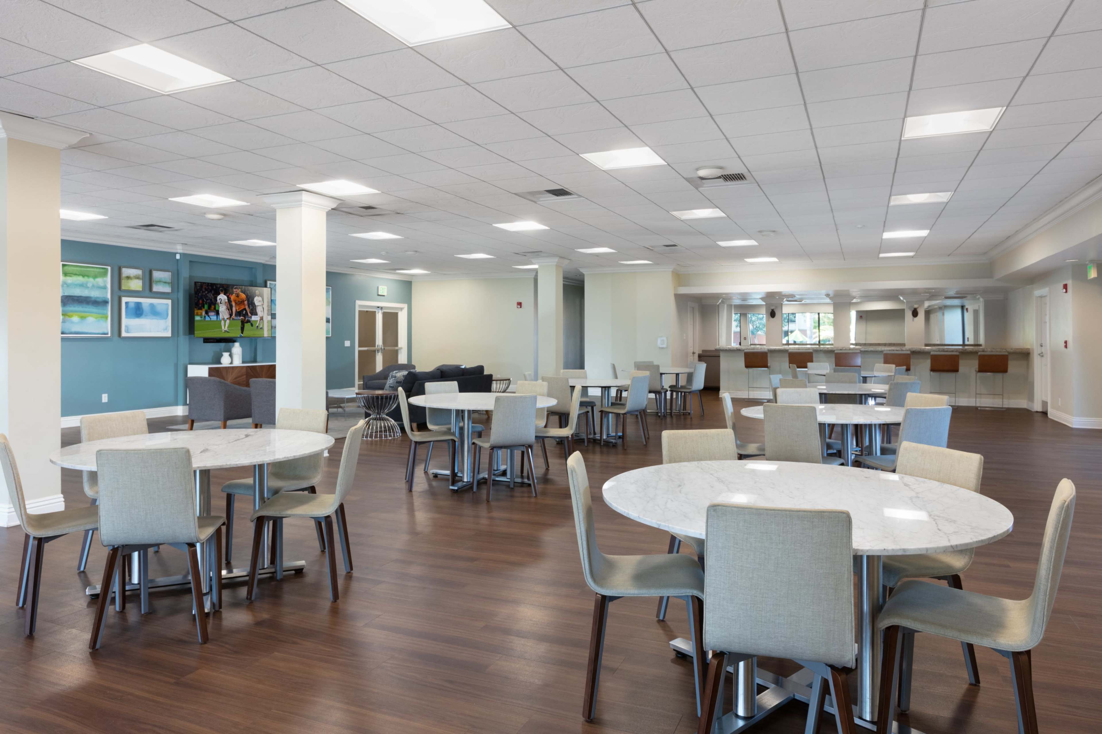 The image shows a spacious communal dining area with round tables and chairs, featuring a counter in the background and large windows allowing natural light.