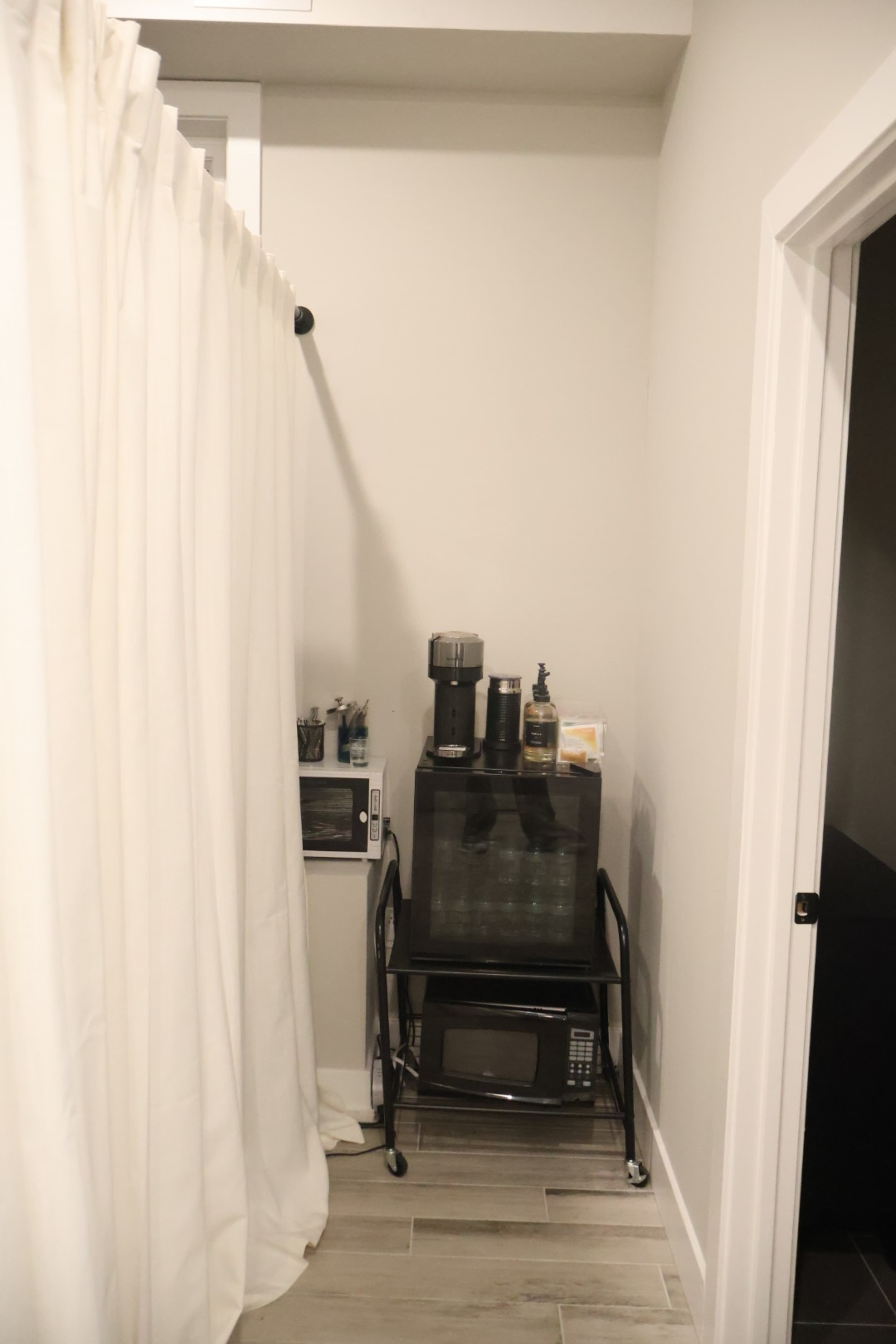 The image shows a narrow corner of a room with a black cart holding a microwave and a coffee maker, next to a curtain that separates the space.