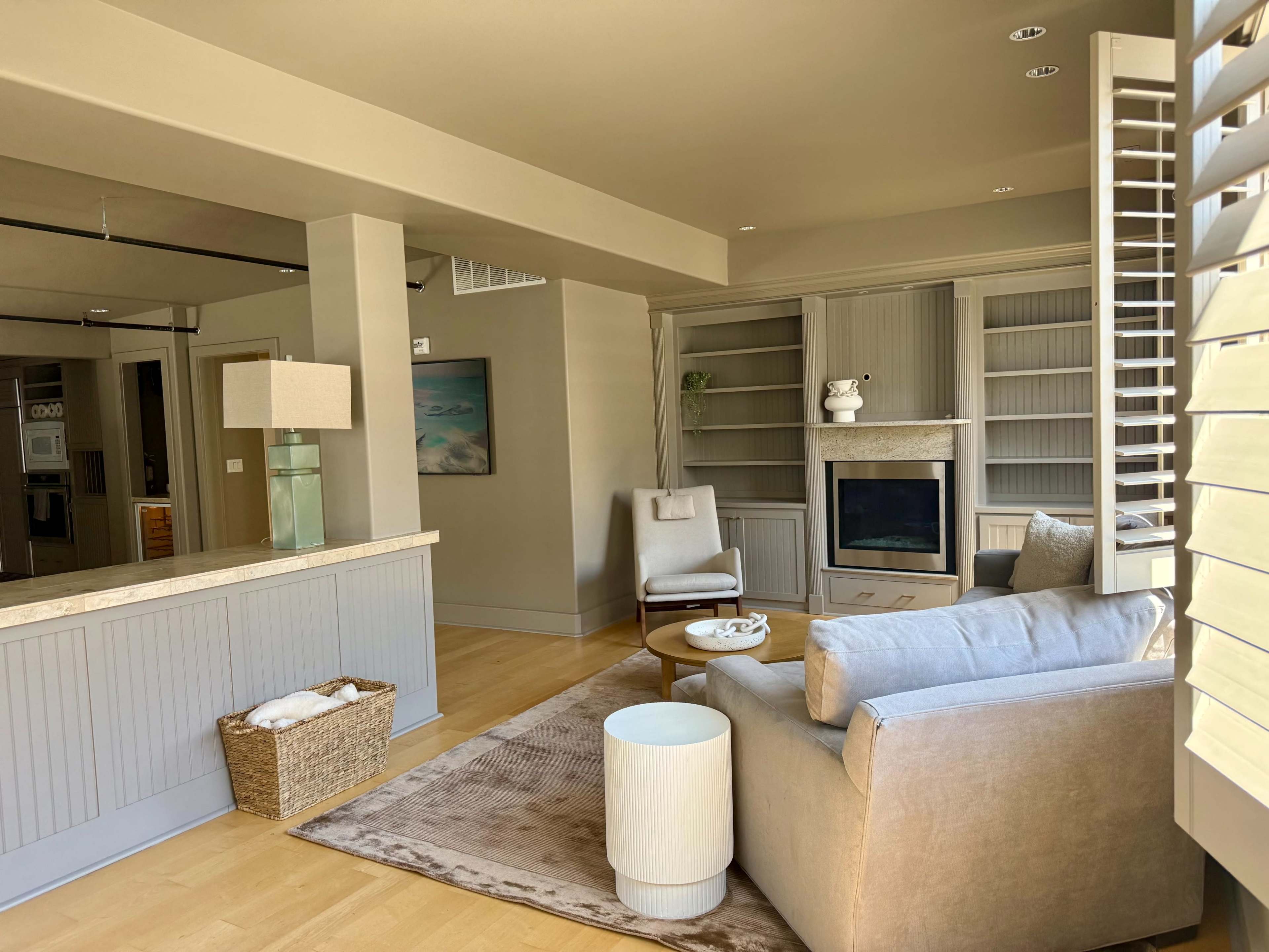 The image shows a modern living room with a light-colored sofa, a round table, an armchair, built-in shelves, and a stone fireplace.