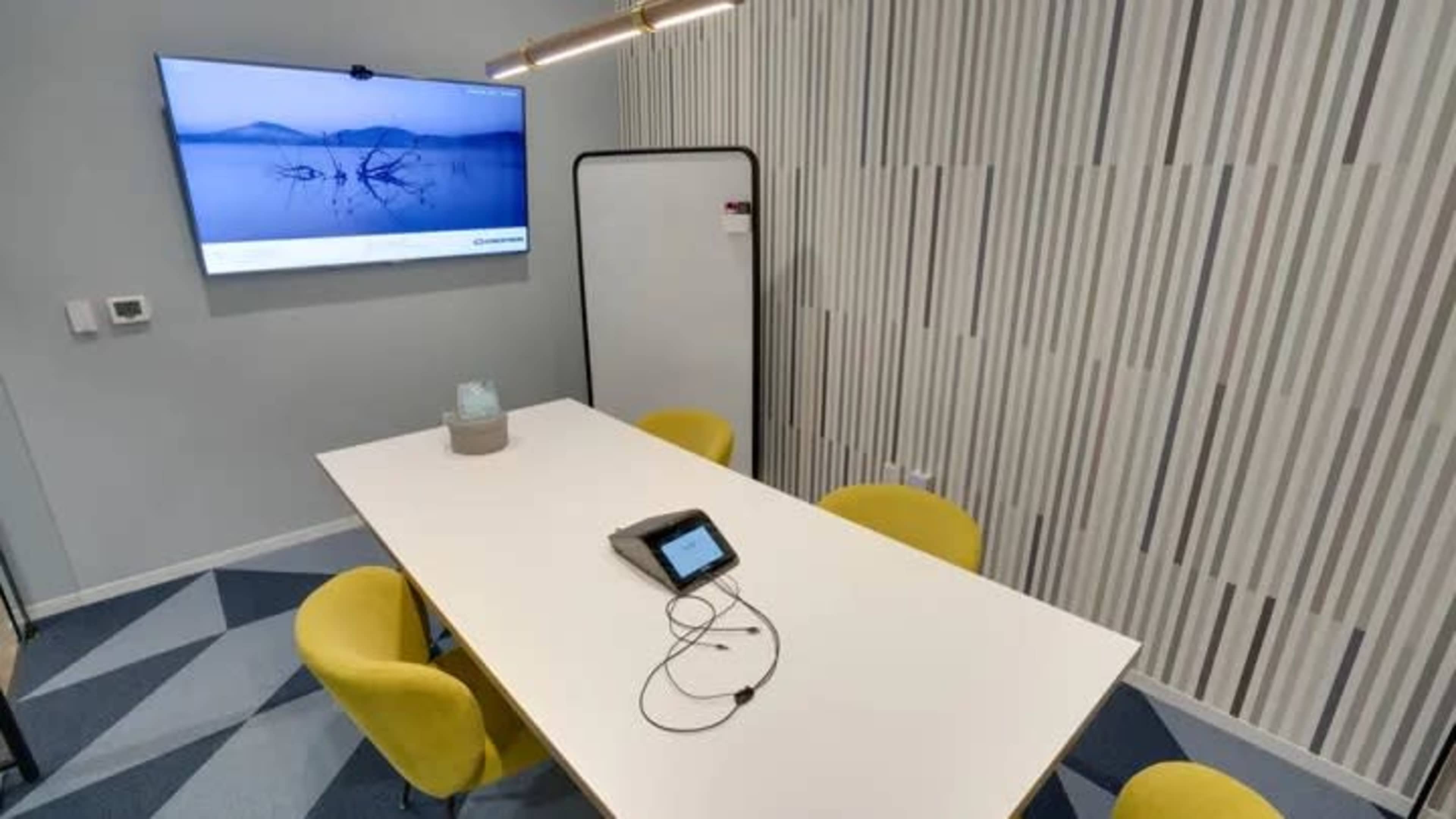 A conference room features a long white table, several yellow chairs, a wall-mounted television displaying a landscape, and a portable whiteboard.